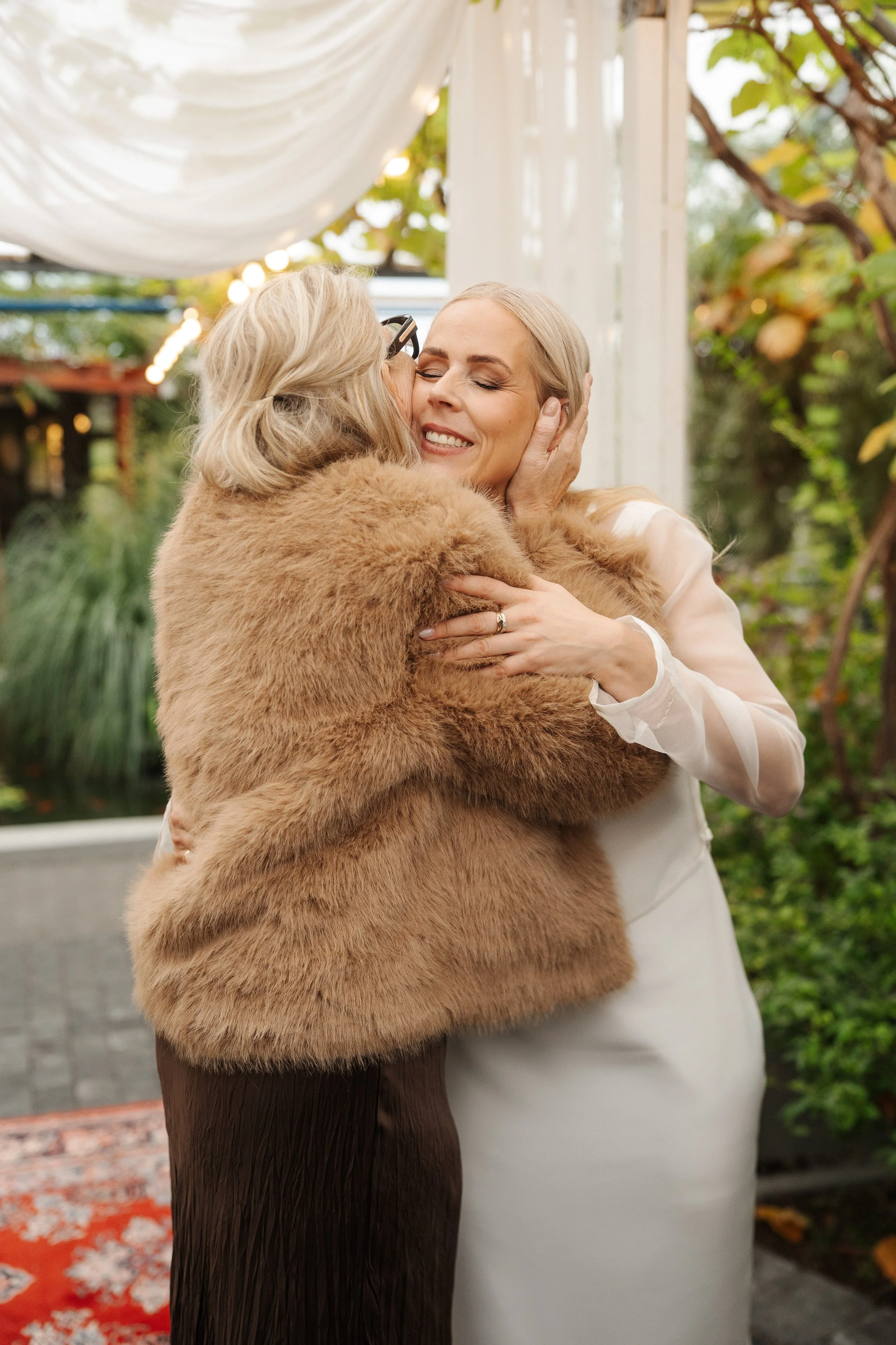 Two women hugging at an outdoor wedding, one in a white dress and the other in a fur coat, smiling with eyes closed.
