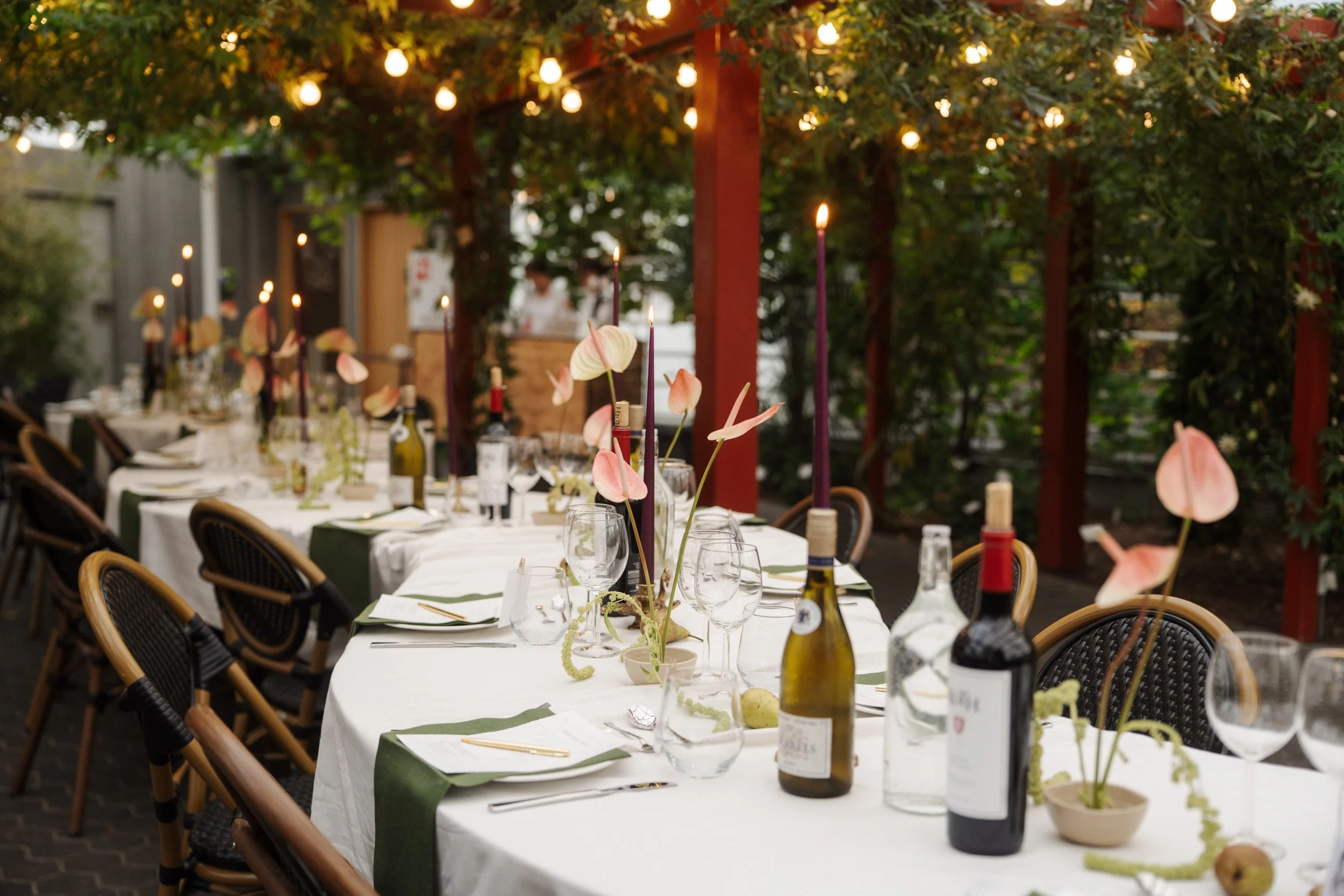 An outdoor dining table set with white tablecloths, wine bottles, glassware, and pink flower centerpieces, illuminated by hanging string lights surrounded by greenery.