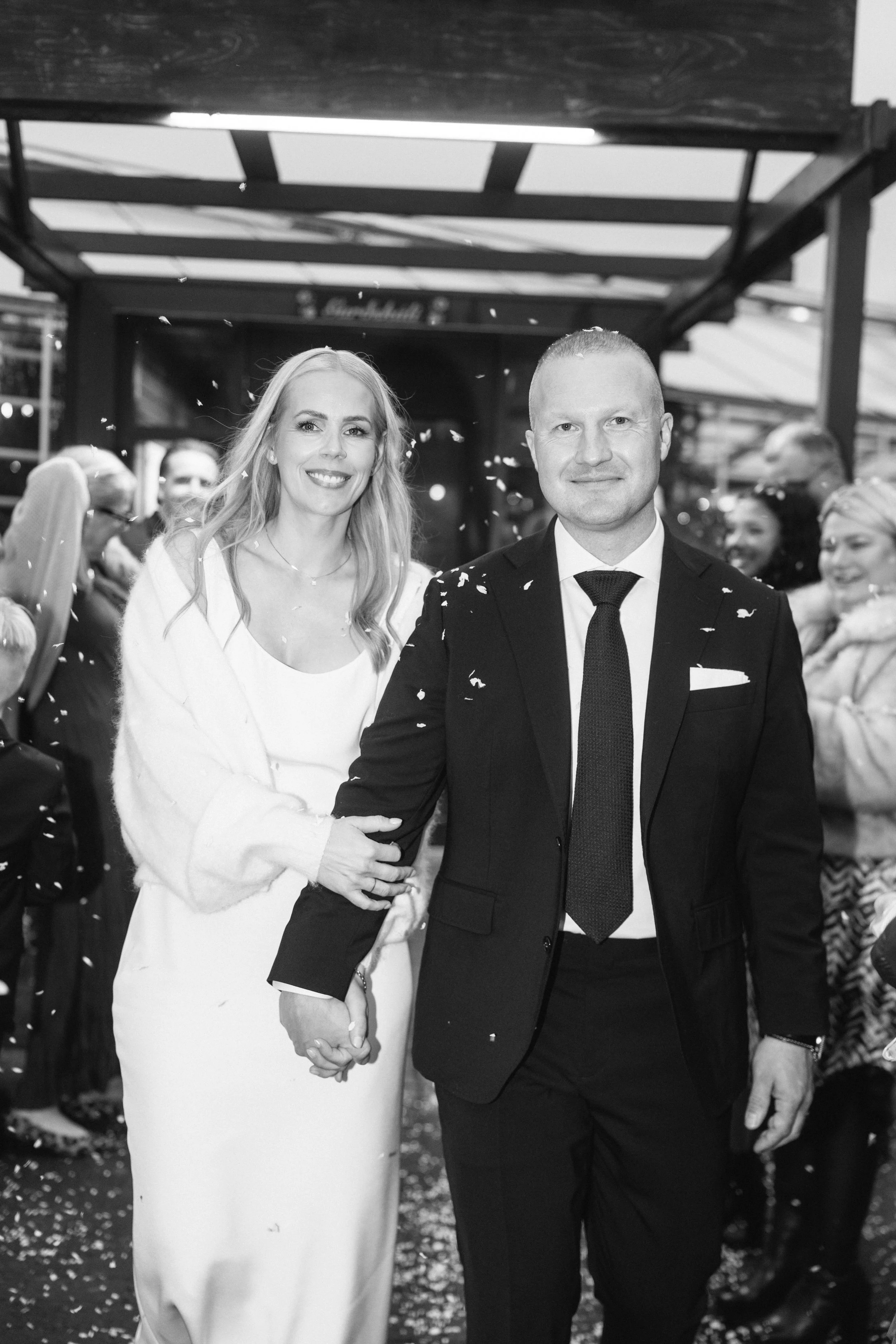 Black and white photo of a couple at a celebration, with the woman holding the man's hand, surrounded by friends and family, with confetti falling.