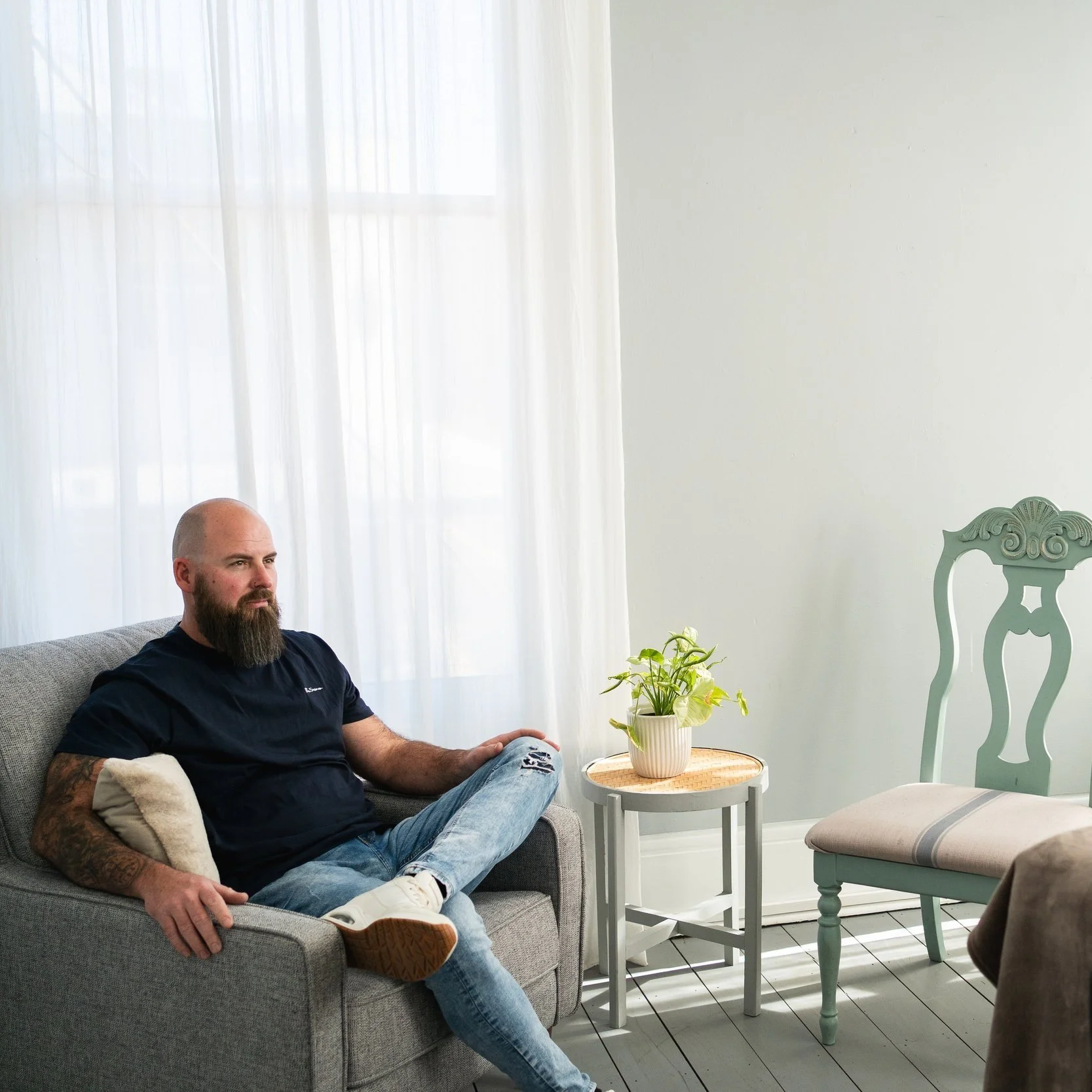 Man with a beard sitting on a gray armchair, wearing a dark shirt and jeans, next to a small round table with a potted plant, in a room with white curtains and a green decorative chair.