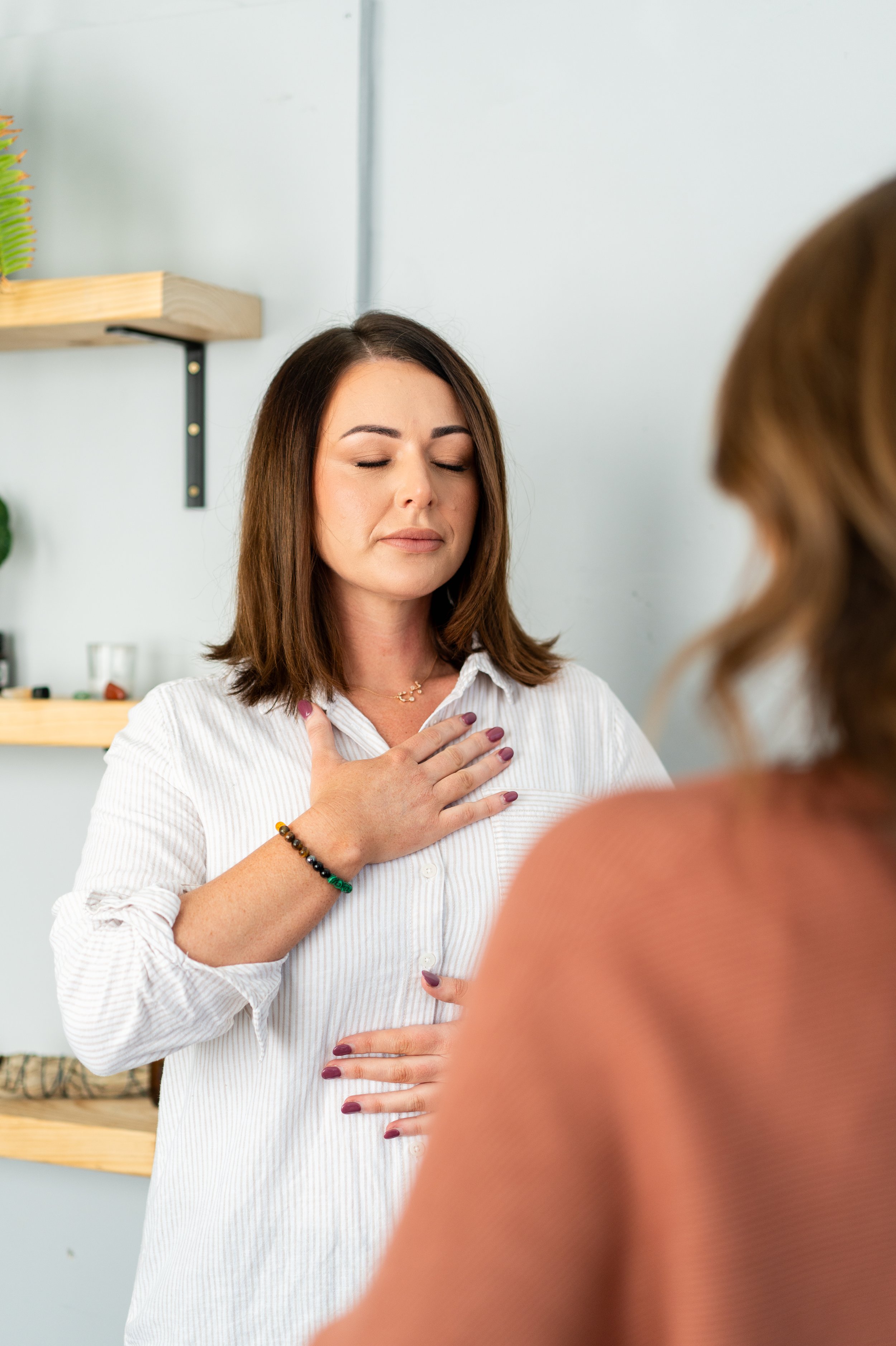 A woman with closed eyes, wearing a white shirt, places her hand on her chest, appearing to be meditating or practicing mindfulness. Another person is partially visible in the foreground.