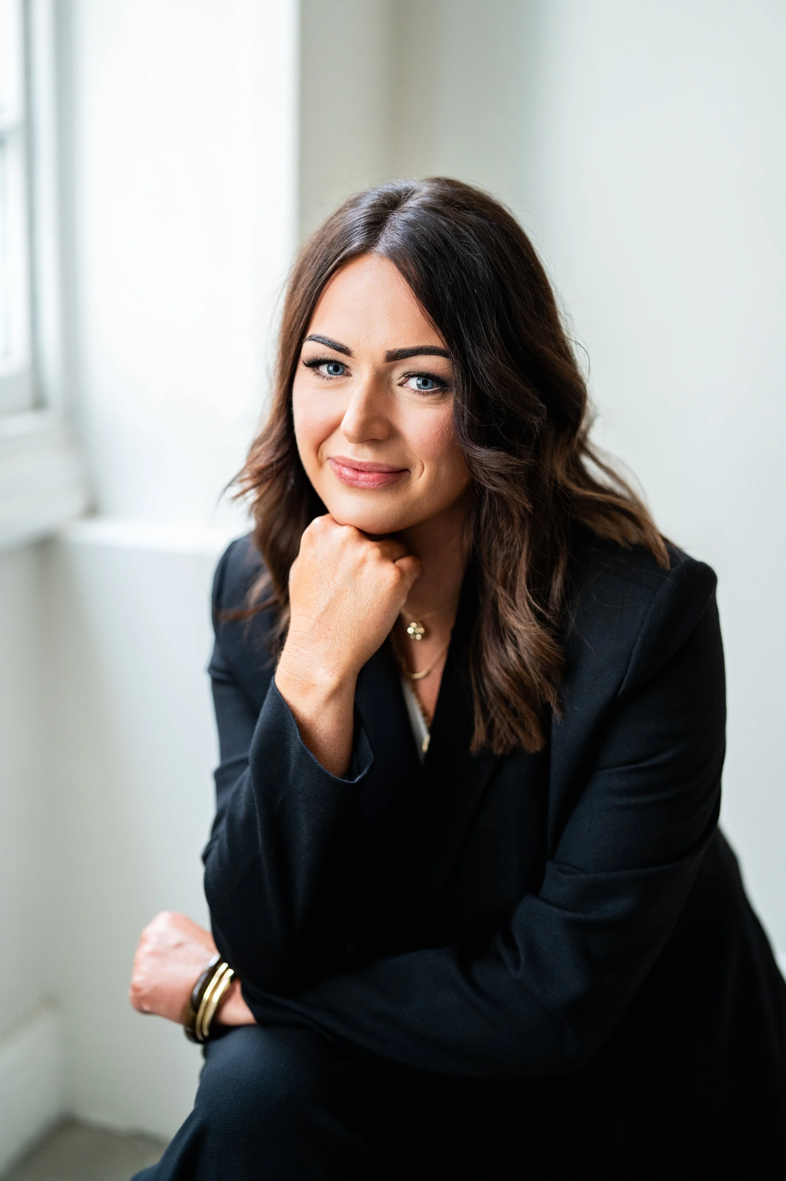 Smiling woman sitting on a sofa, wearing a black top and light pants, accessorized with hoop earrings and bracelets.