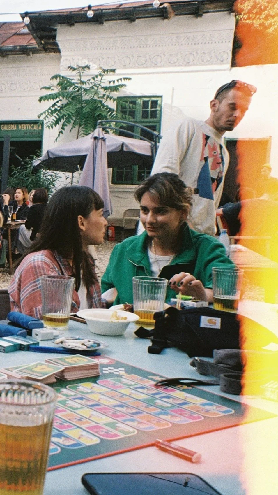 Two women sitting at a table playing a card game outdoors, with drinks and snacks on the table, while a man stands nearby. In the background, other people are seated at tables, under a patio umbrella, with a building that has decorative molding and leafy plants.