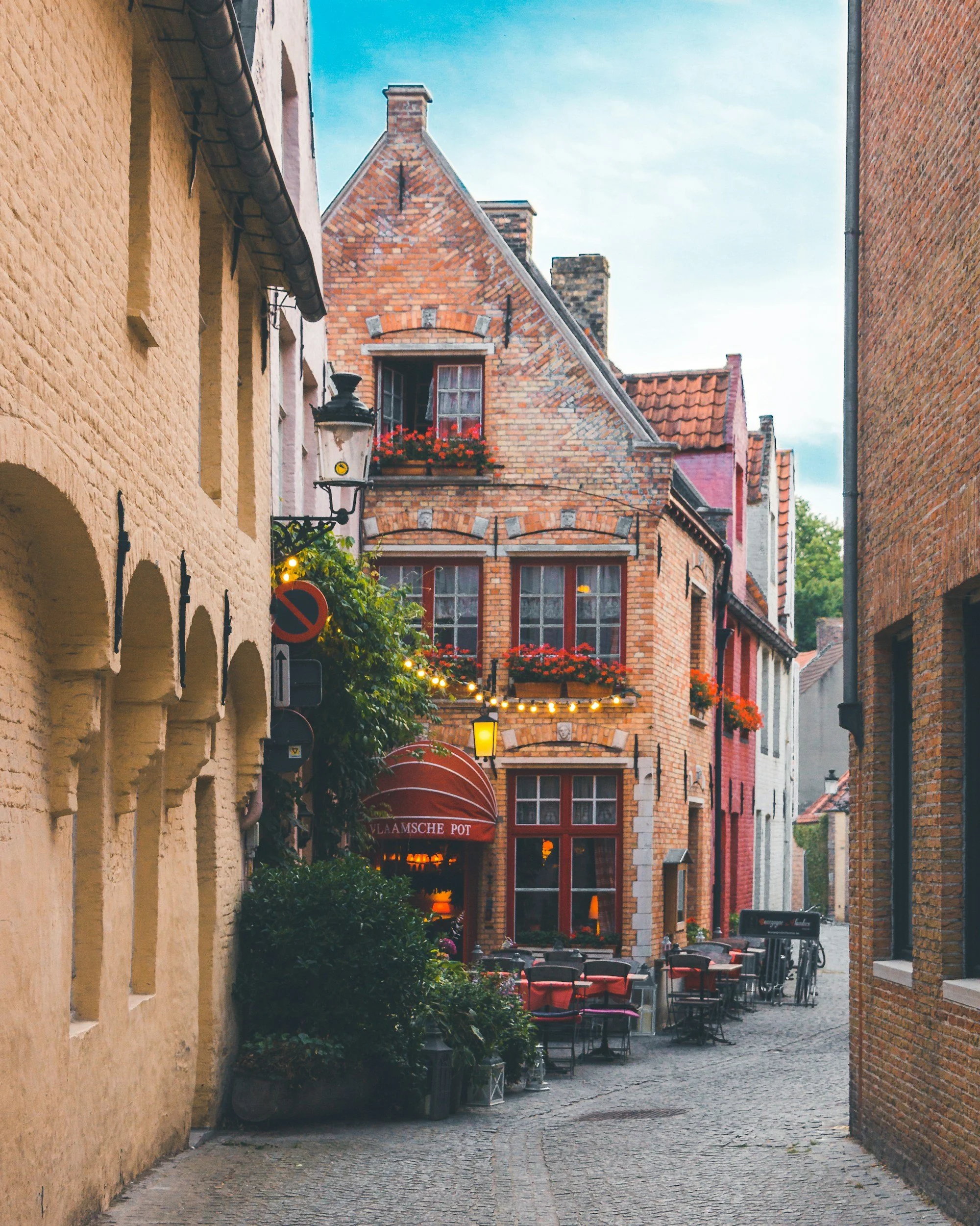 A narrow cobblestone street in a European city with colorful brick buildings, outdoor cafe seating, string lights, and flower boxes under the windows.