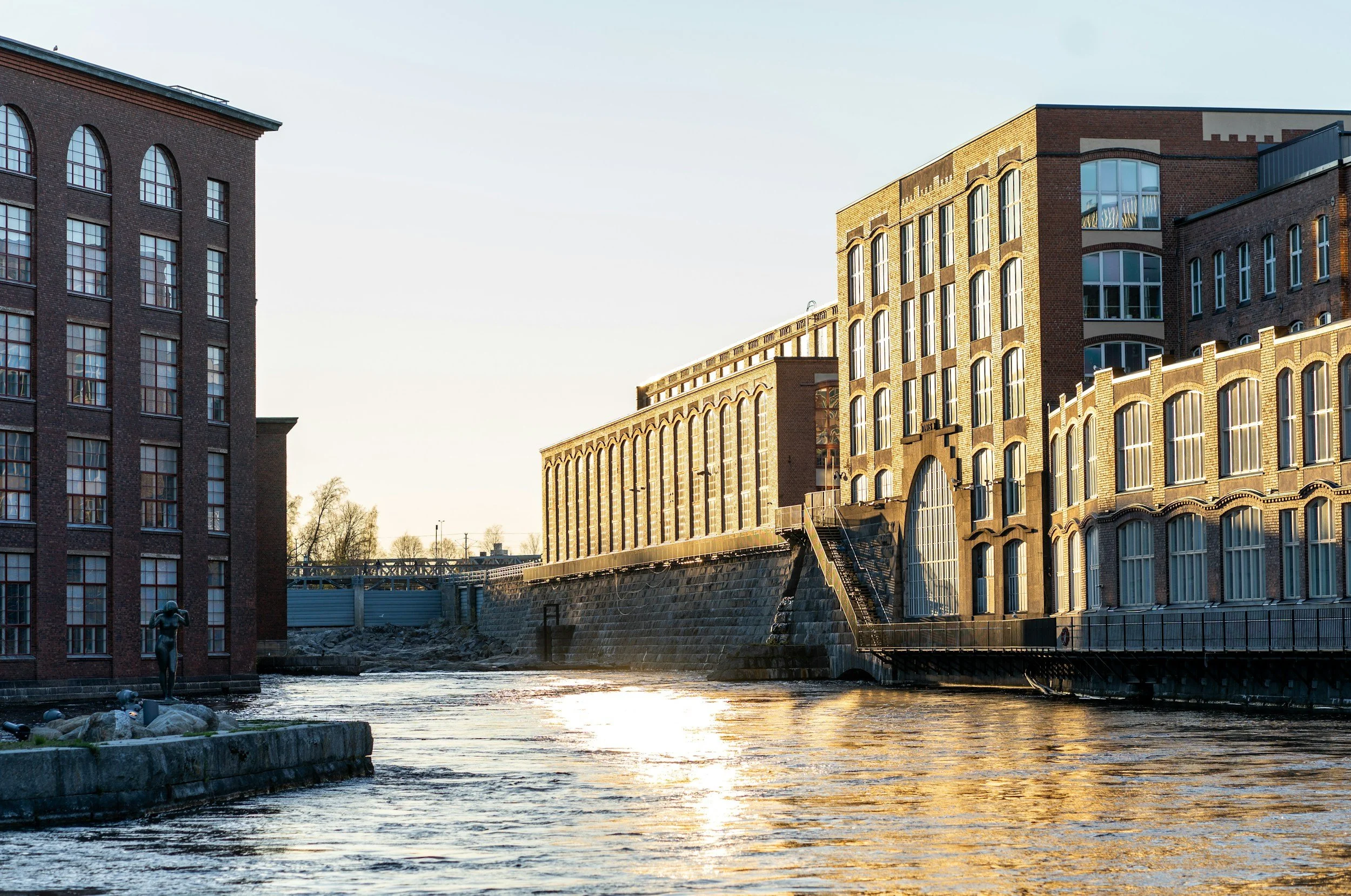 Sunset view of historic brick buildings along a river, with a statue on the riverbank in the foreground.