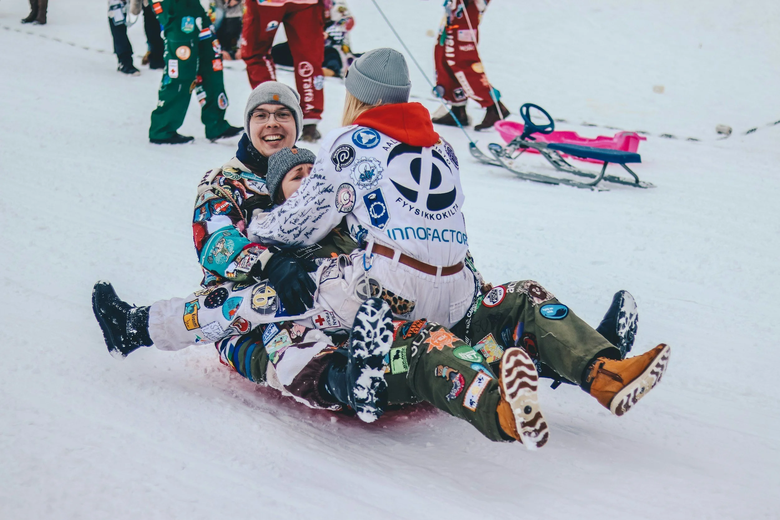 Three children in colorful winter clothes sitting and lying on a sled on snow, surrounded by other children and sleds in the background.