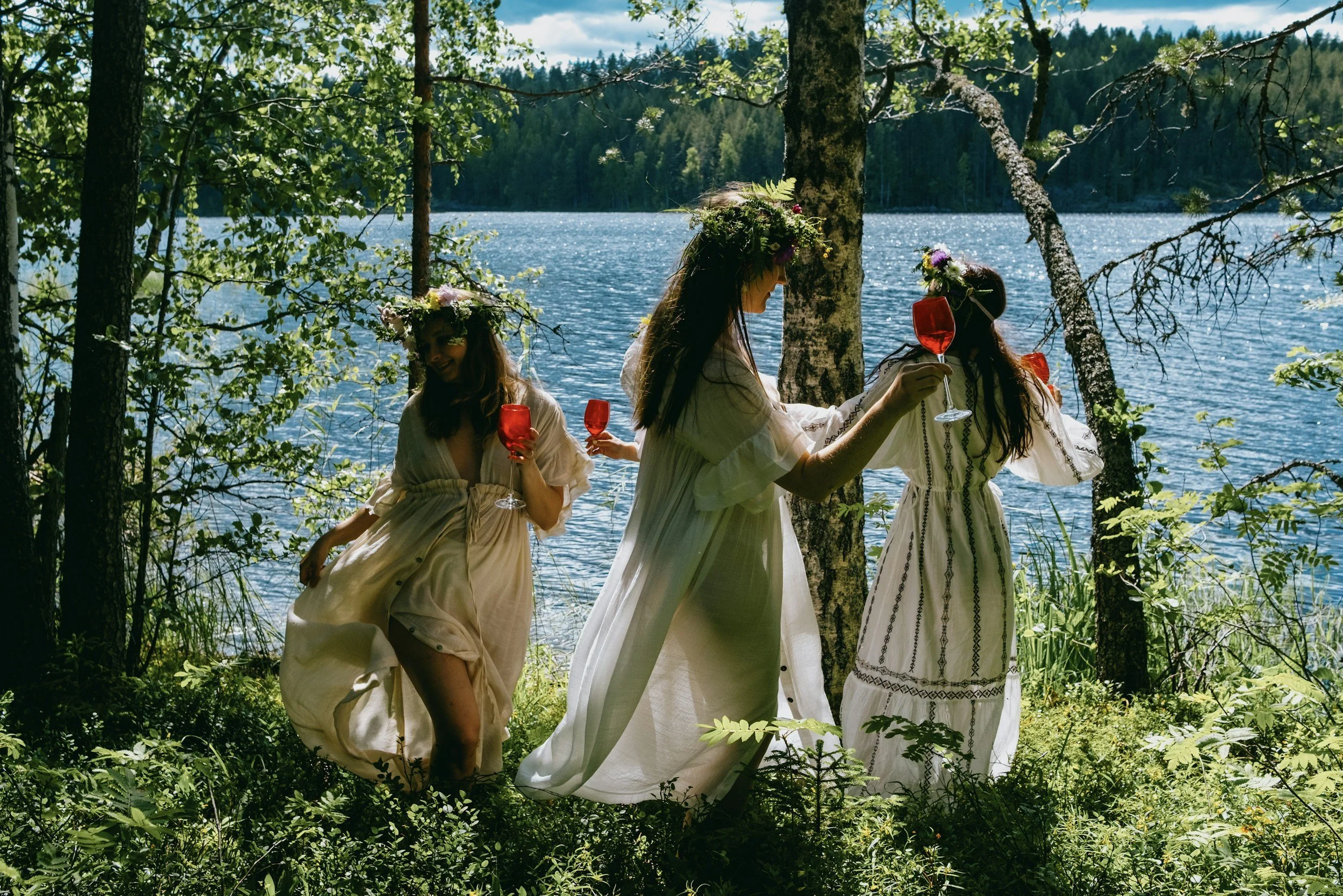Three women in white dresses with flower crowns holding red glasses by a lakeside in the woods.