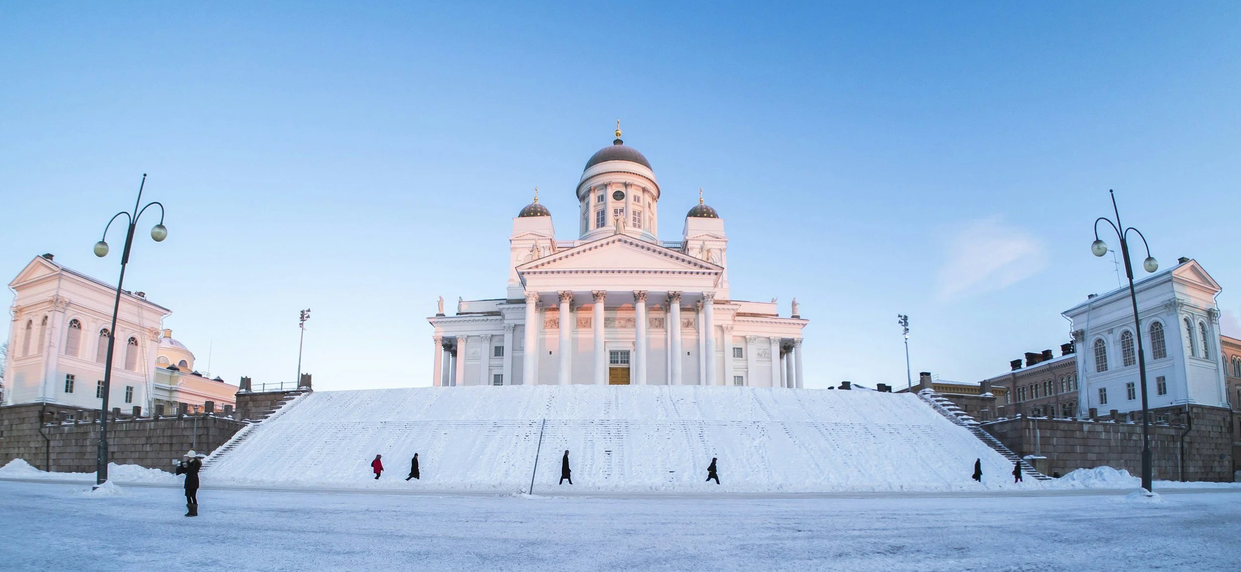 A large white church with a central dome and four smaller domes, set against a clear blue sky, with snow-covered ground and several people walking by.