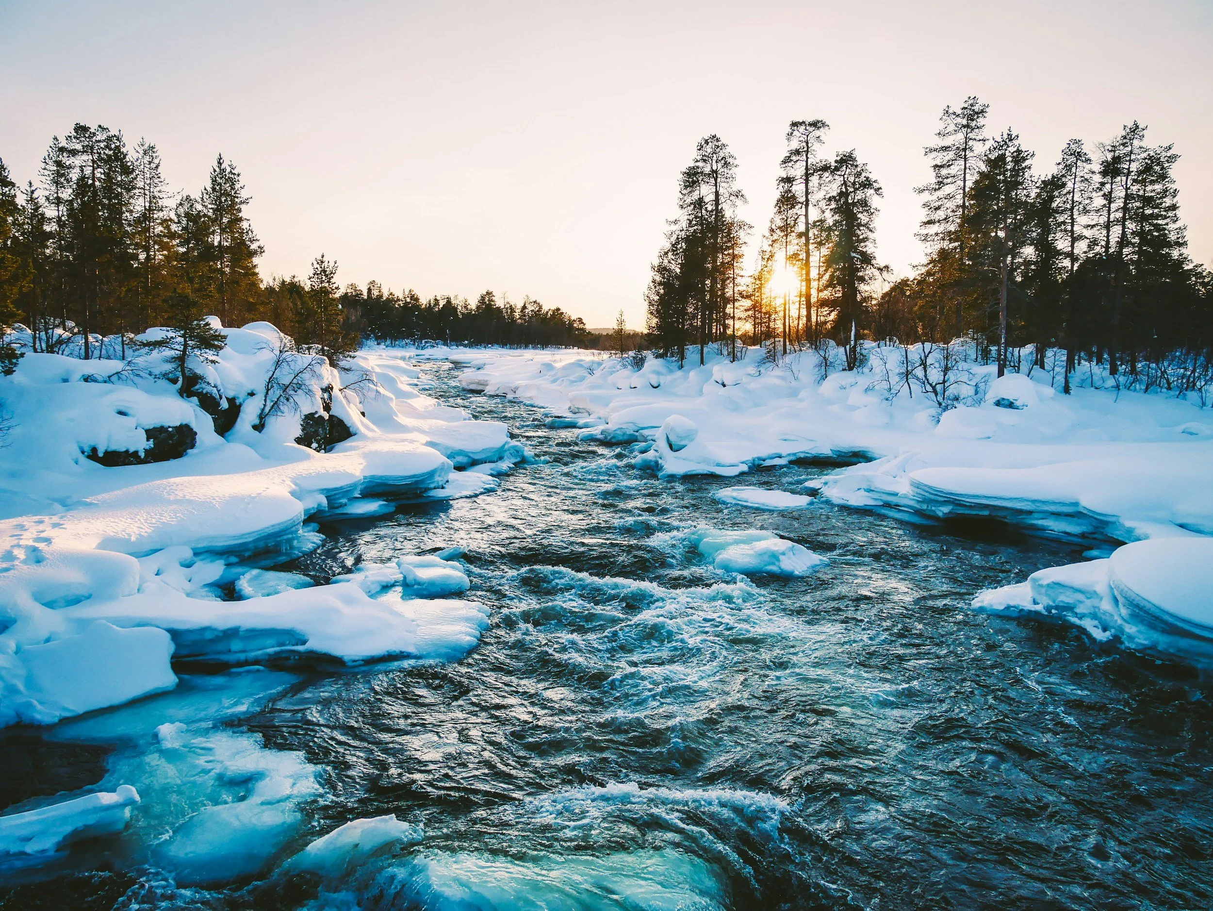 Snow-covered river flowing through a winter forest at sunset.