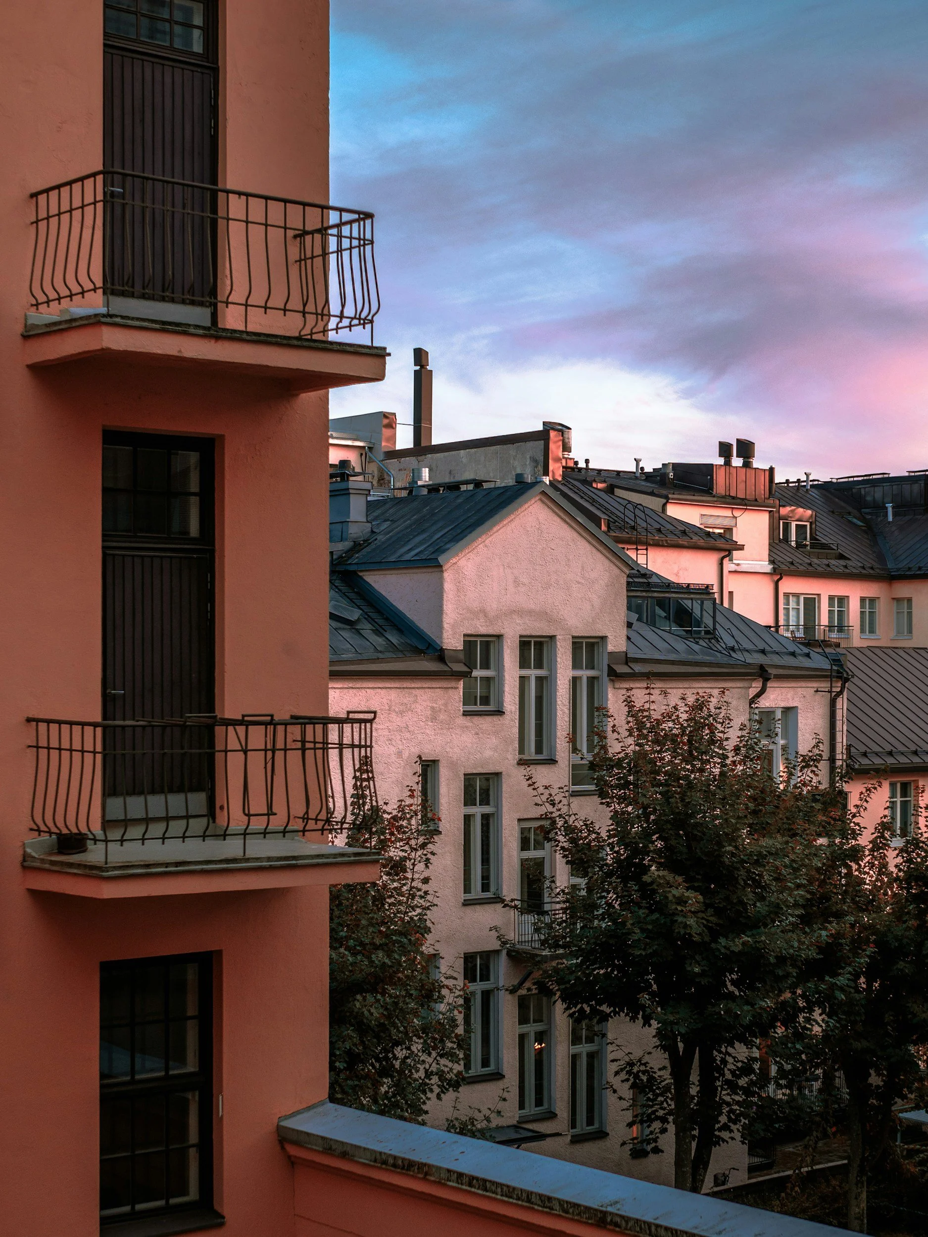 View of pink and beige residential buildings with dark metal roofs and balconies, trees, and a colorful sky at sunset.