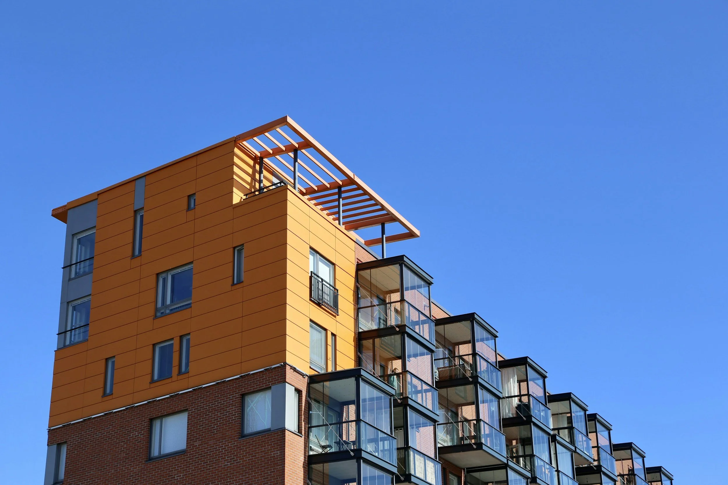 Modern multi-story apartment building with orange and red exterior, featuring glass balconies and a rooftop structure, against a clear blue sky.