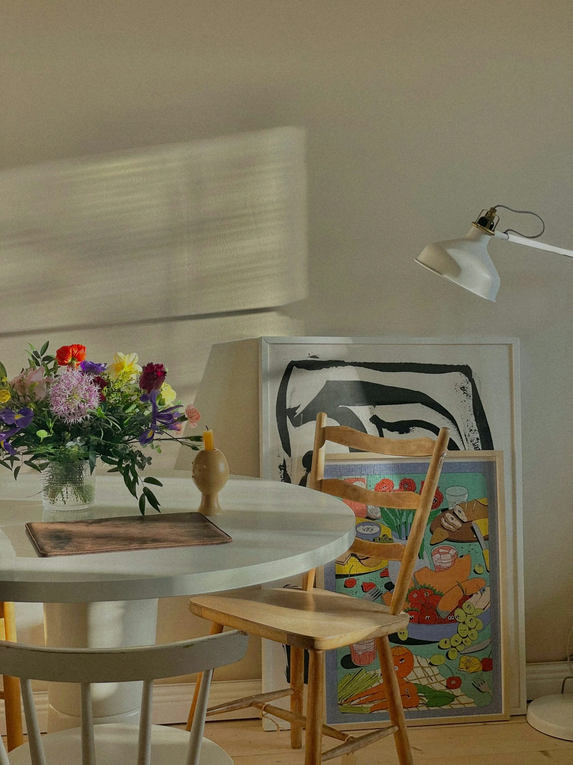 Interior scene with a white round table holding a bouquet of colorful flowers and a small tan vase, surrounded by wooden chairs. Several framed artworks are stacked behind the chairs, with a white wall and a white adjustable lamp overhead casting a shadow on the wall.