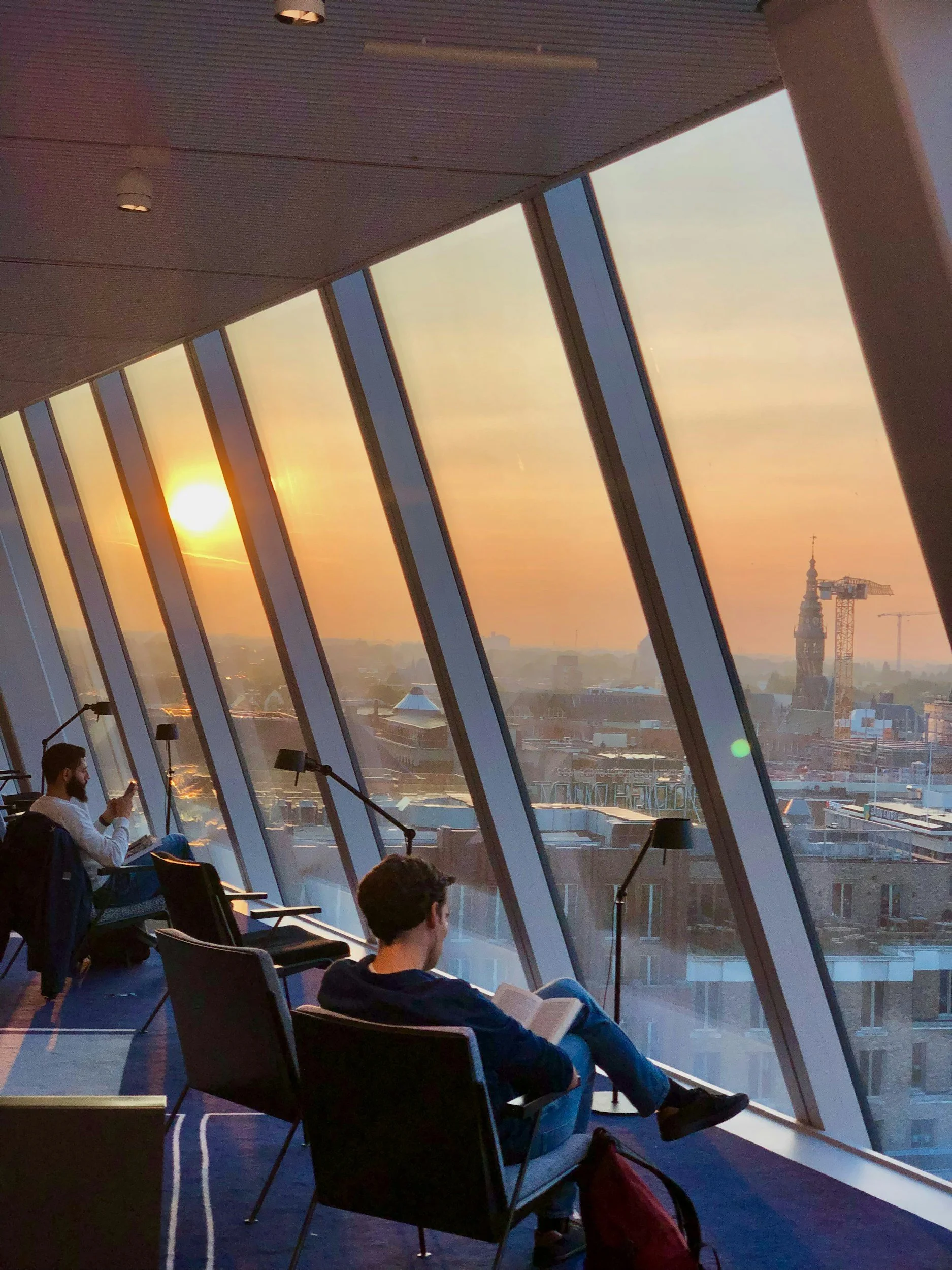 Two people sitting in a modern, slanted window view room during sunset, one reading a book and the other using a smartphone, with city buildings and cranes outside.