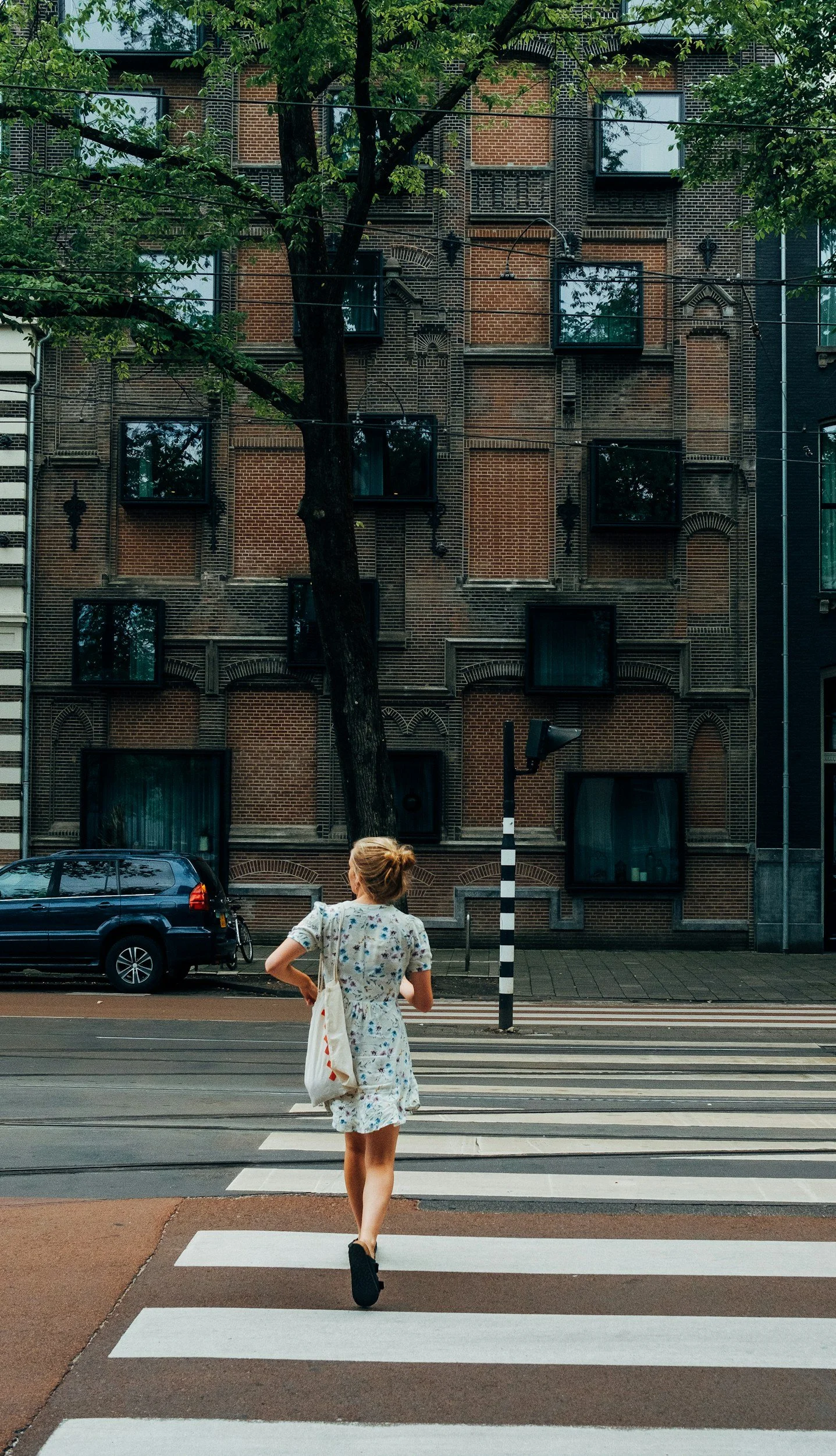 A woman in a floral dress and black shoes walking across a pedestrian crosswalk in front of a brown brick building with multiple windows, a large tree, a parked car, and street lamps.