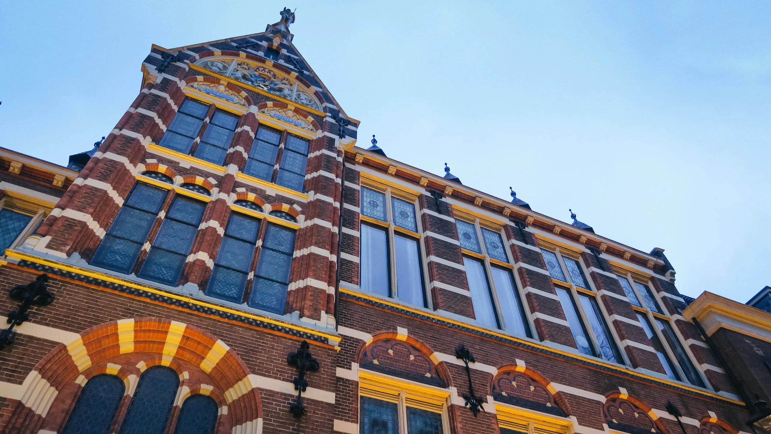 Low-angle view of a historic brick building with decorative window trim and a tower with a weather vane, set against a partly cloudy sky.