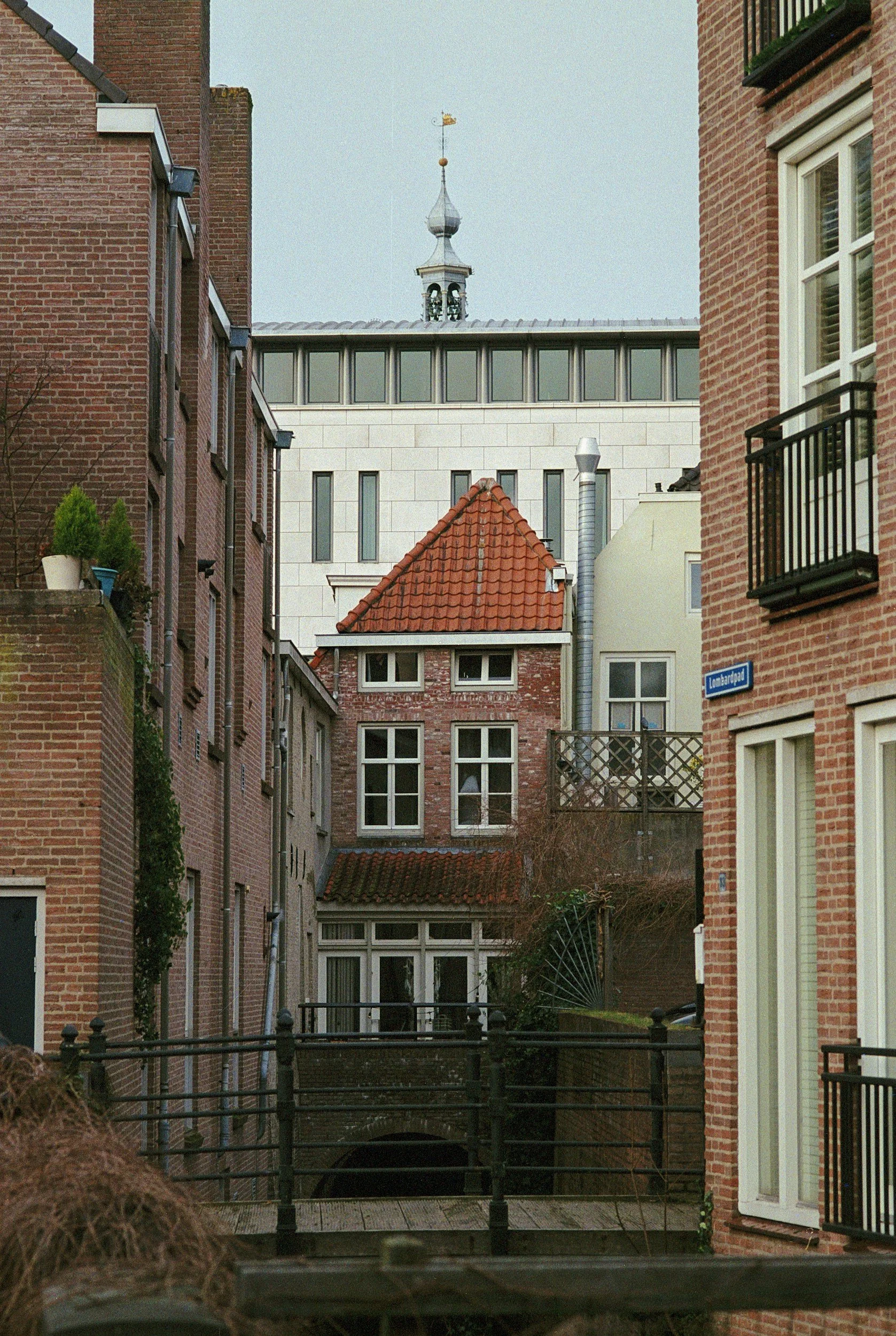 View of old brick residential buildings with small windows, balconies, and red tiled roofs, with a modern white building and a tower in the background.