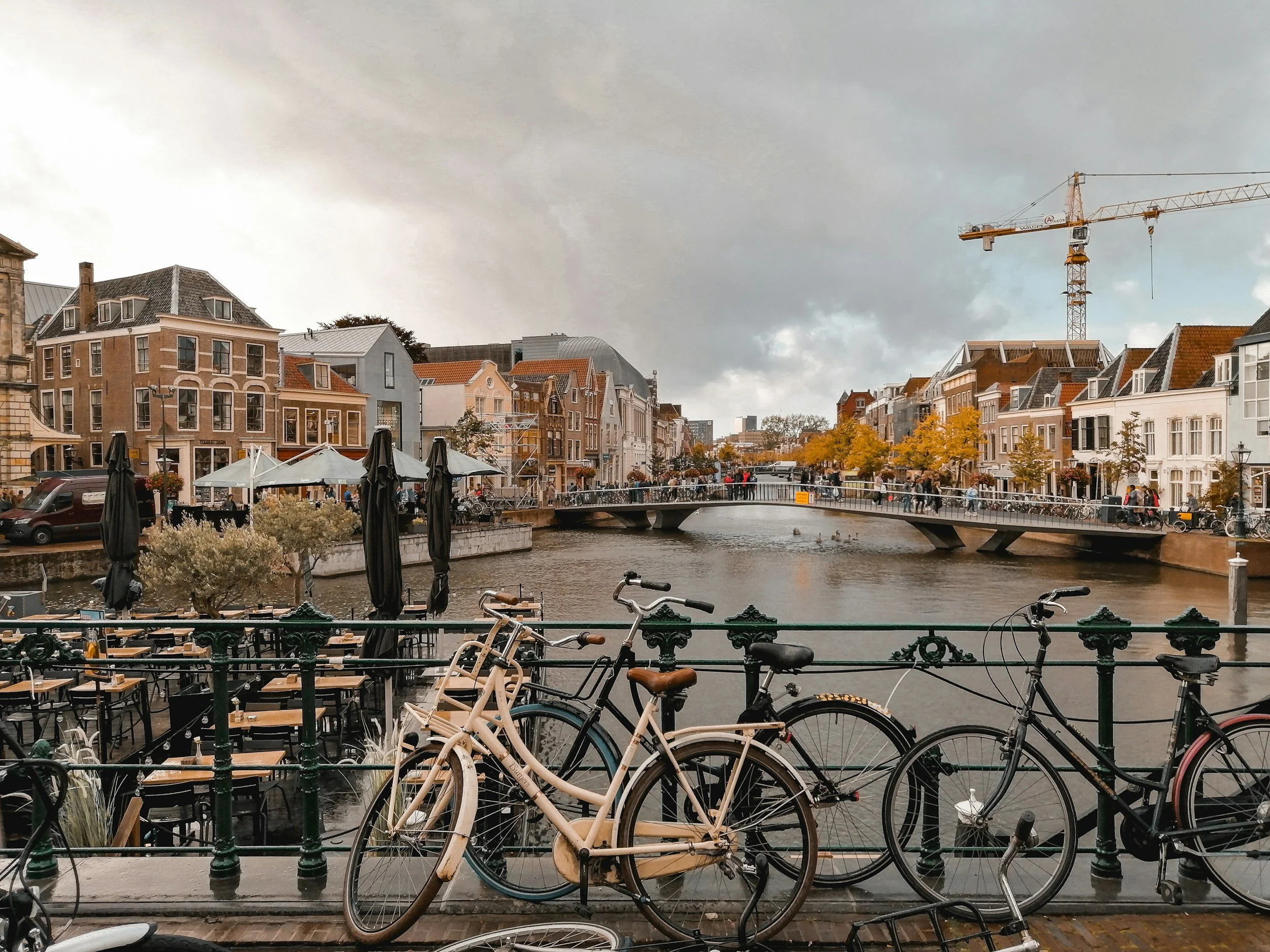 Bicycles parked along a railing overlooking a canal with a bridge, lined with colorful buildings, trees, and outdoor cafes under a cloudy sky.
