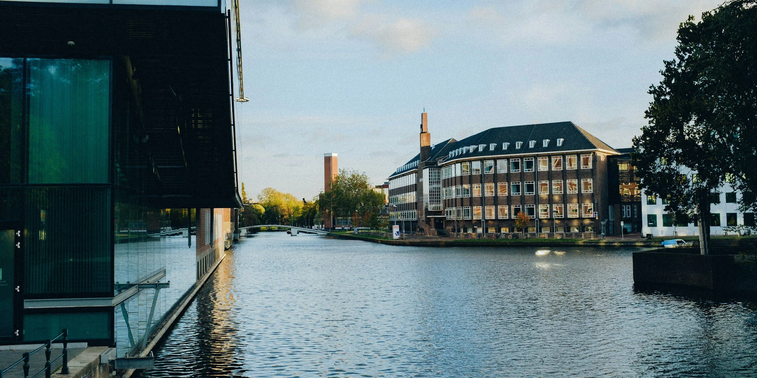 A view of a canal with modern buildings reflecting in the water, trees, and a boat moving on the water, under a partly cloudy sky.