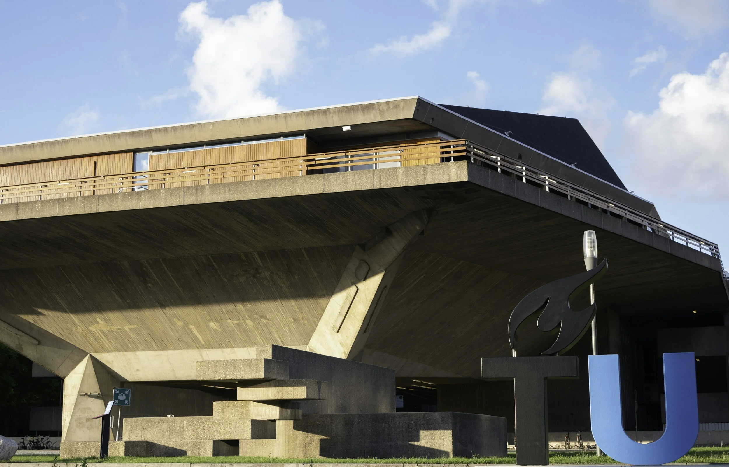 Modern university building with concrete overhang, metal and wood railing, large Danish U statue in front, bicycle racks, and a blue sky with clouds.
