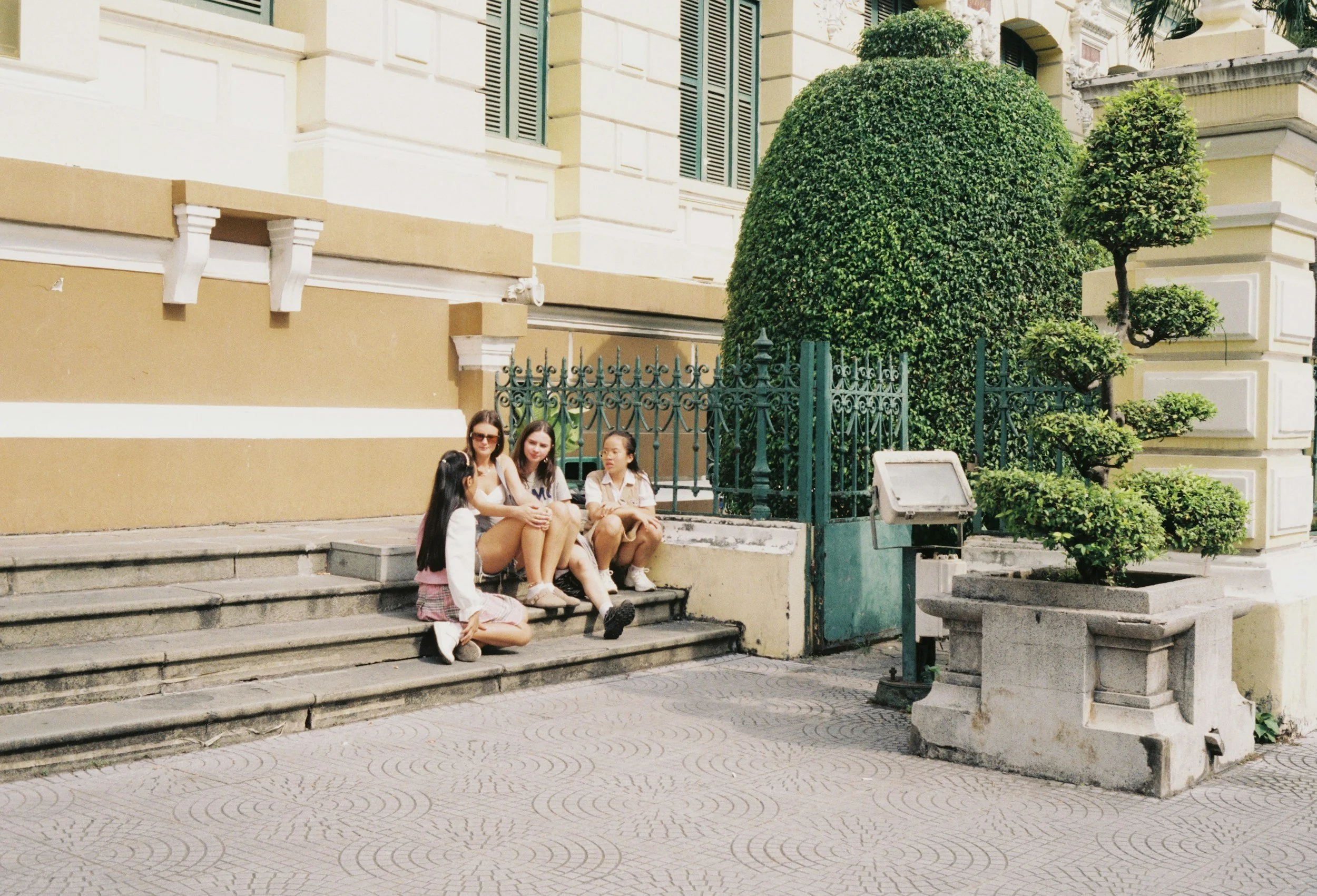 Four young women sitting on steps outside a building with a yellow facade, green gate, large trimmed bushes, and decorative plants.