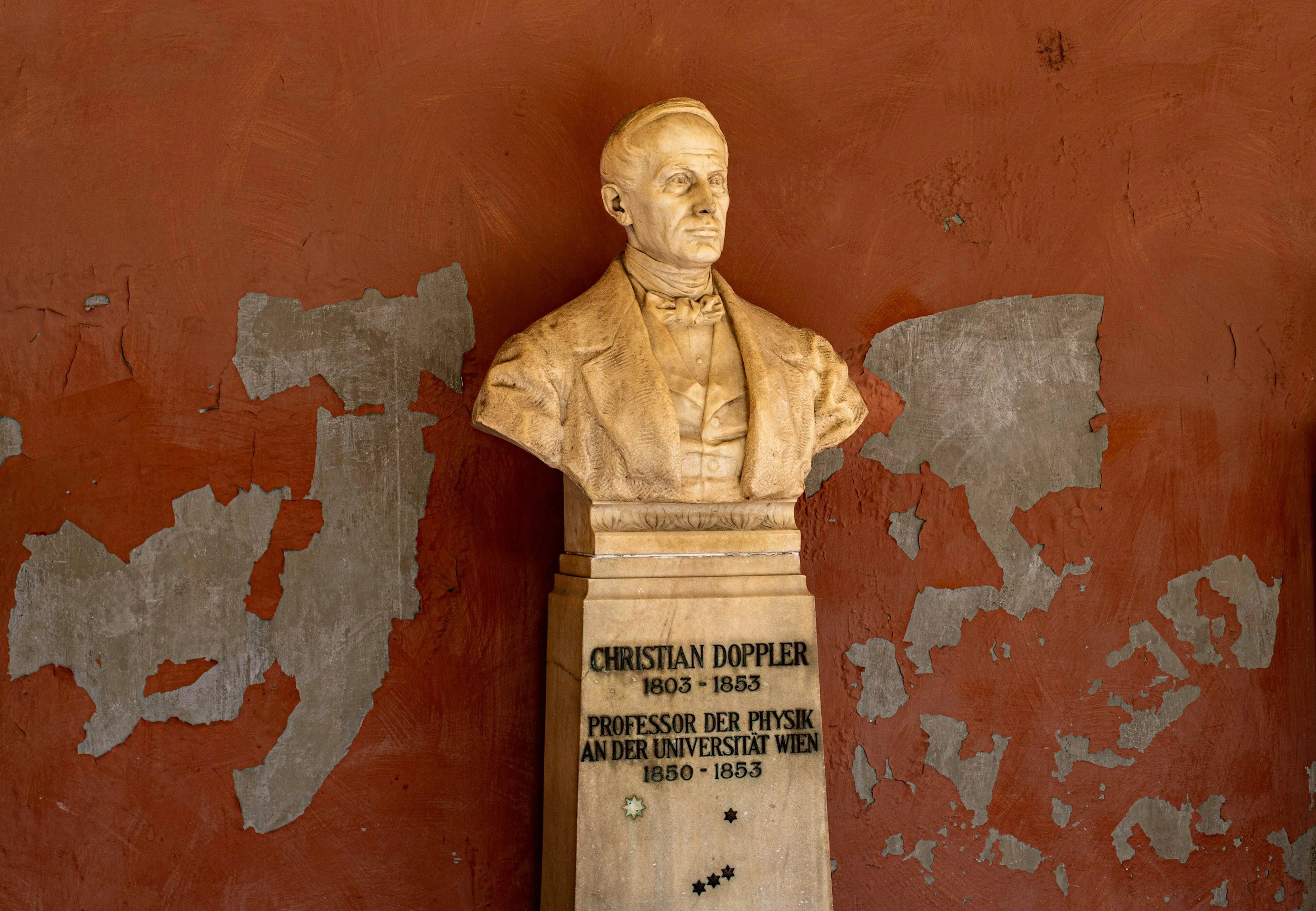 Bust of Christian Doppler on a pedestal with his name, birth and death years, and his titles written in German, in front of a weathered red wall.