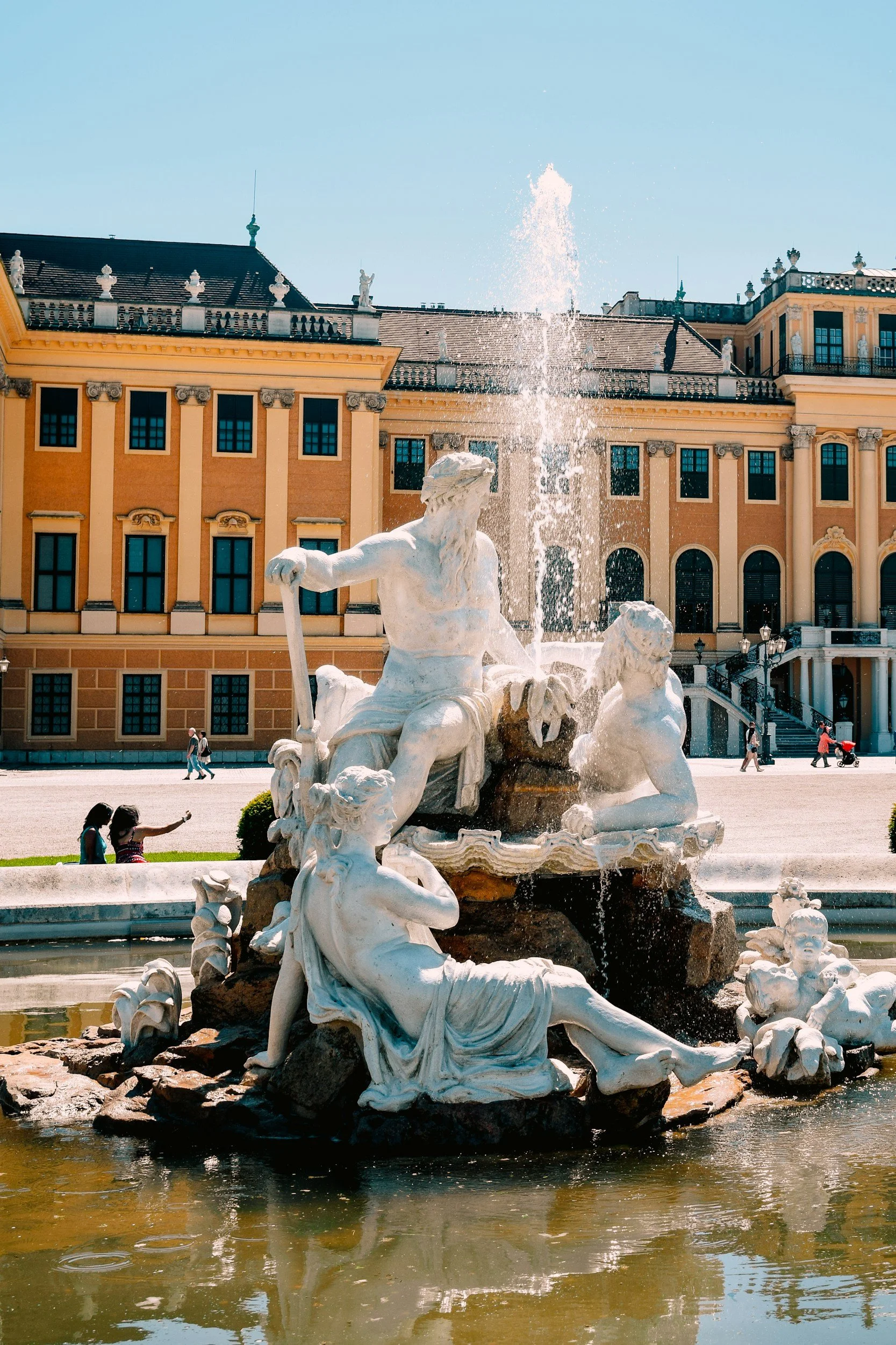 A large ornate fountain with classical sculptures of mythological figures, with water shooting up from the center. In the background, a grand yellow building with many windows and decorative architectural details. There are people walking and sitting around the fountain on a sunny day.