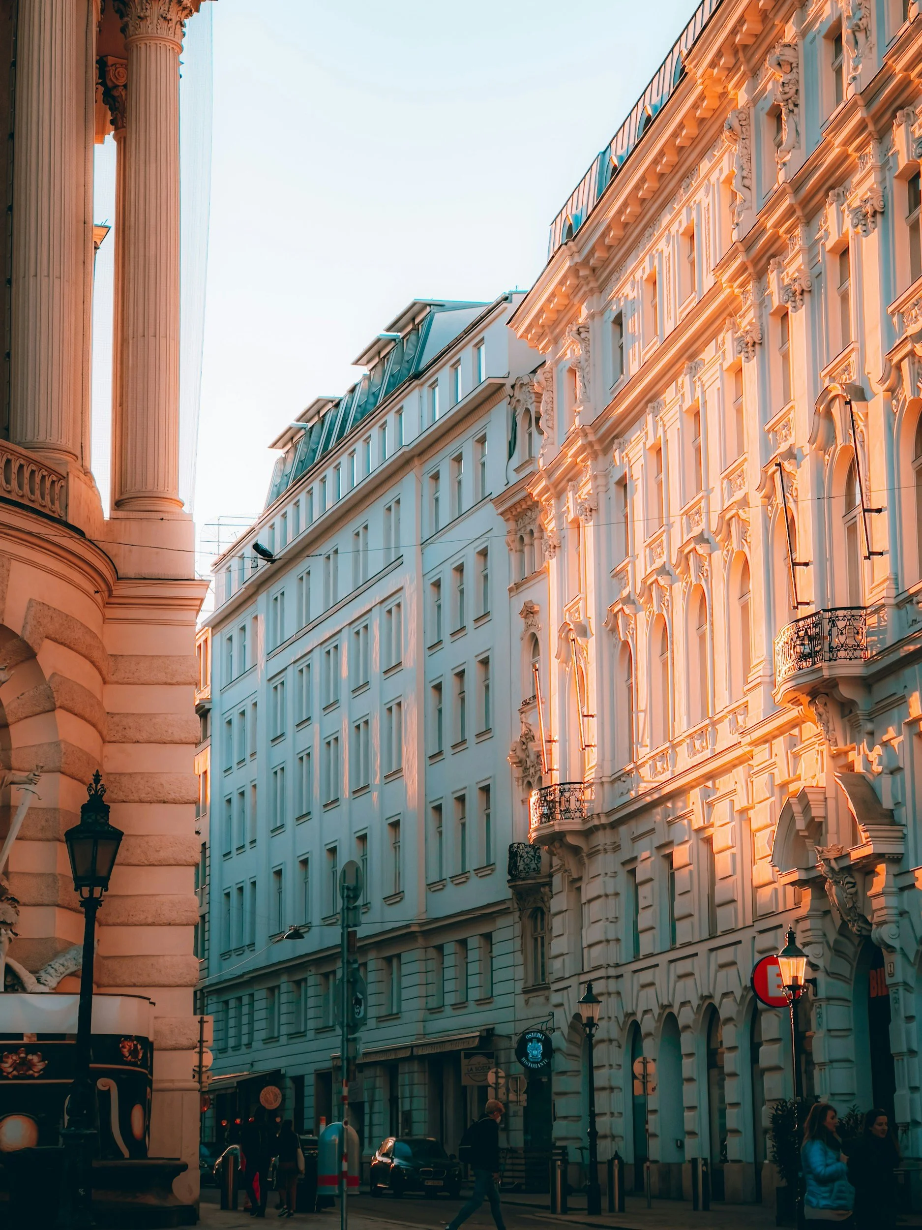 City street with elegant historic buildings illuminated by sunlight, pedestrians walking, street lamps, and parked cars.