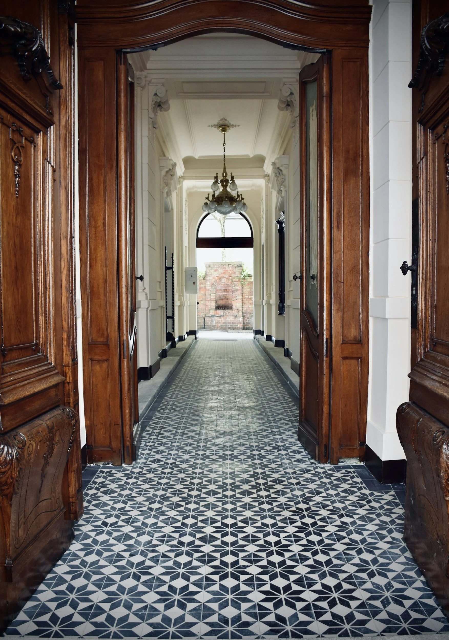 A vintage entrance hallway with patterned black and white tile floor, wooden door frames, ornate white walls, glass chandelier, and a view of a brick wall outside.
