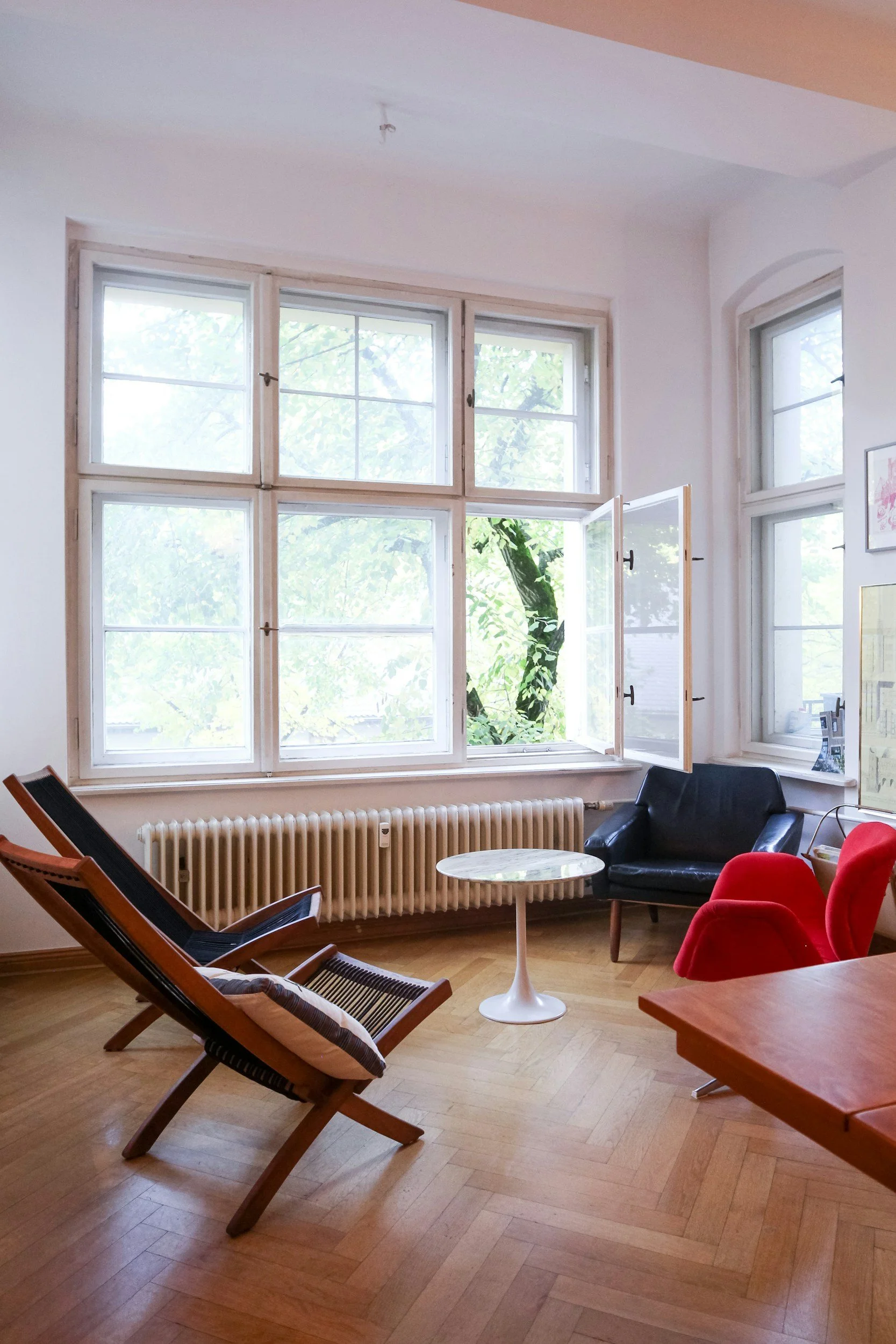 A cozy living room with large windows, a black armchair, two red chairs, a wooden chair with a pillow, a small round table, and a radiator under the windows.