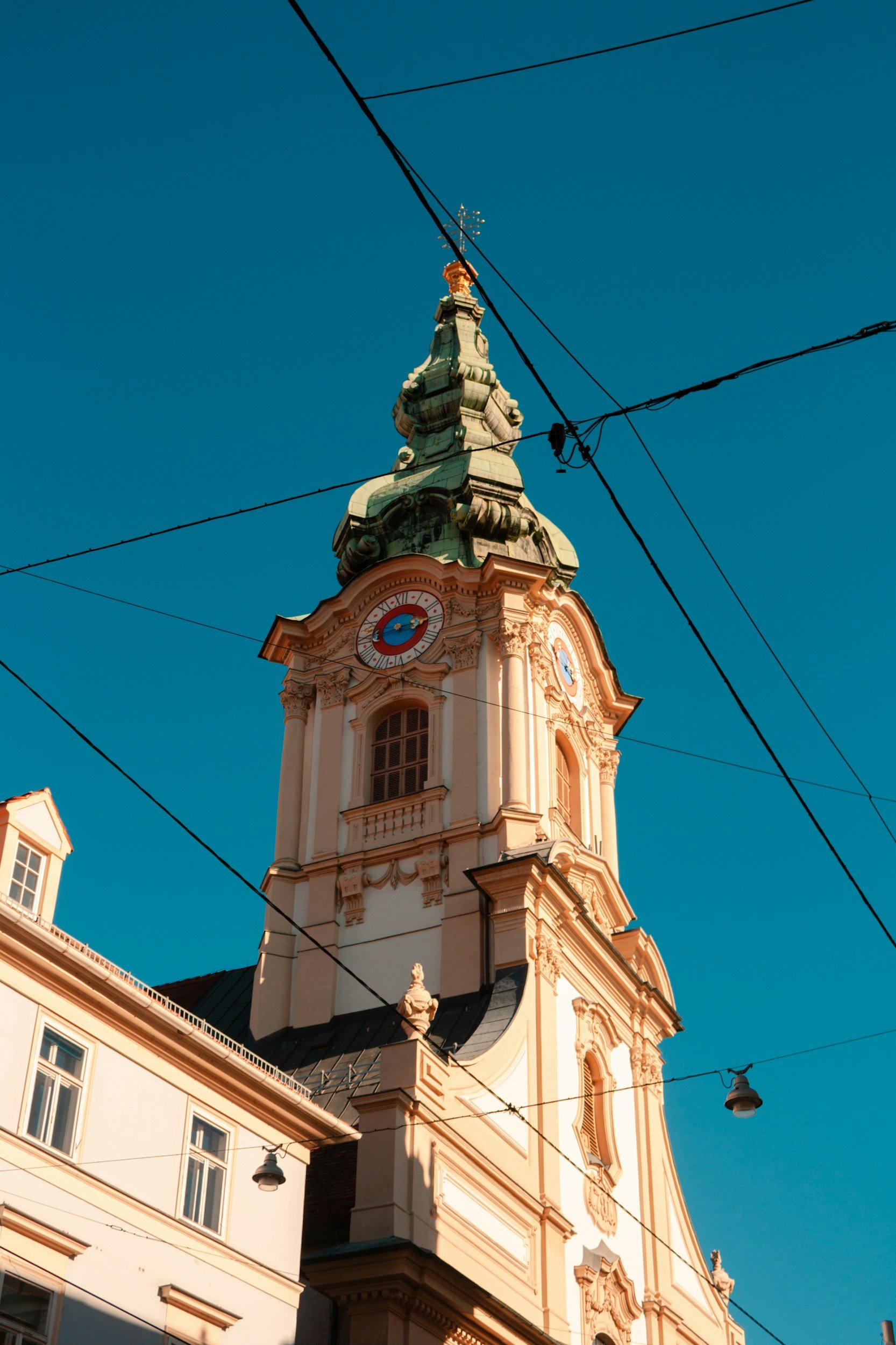 A church tower with a clock, ornate architecture, and a greenish roof, under a clear blue sky with overhead wires.