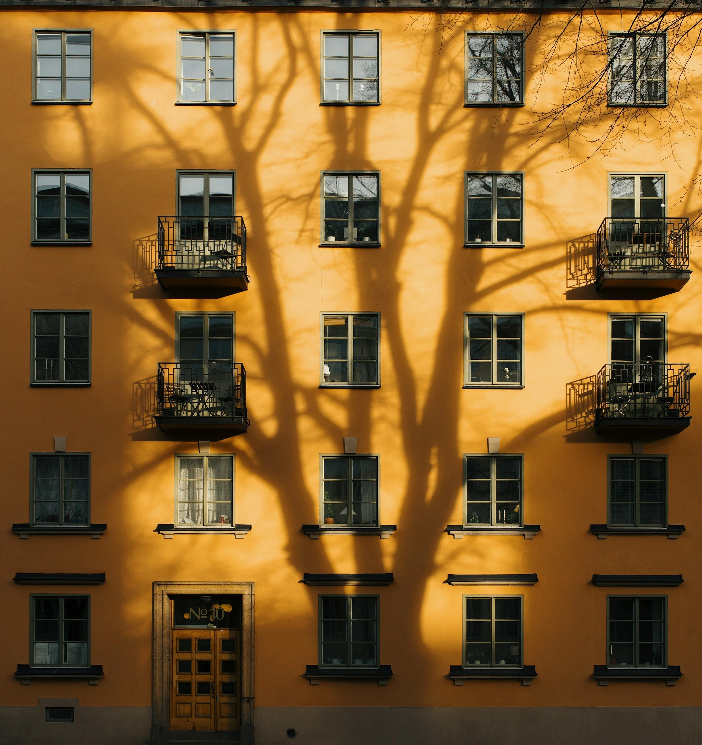 Yellow building with multiple windows and three small balconies. A large tree's shadow is cast on the building's facade.