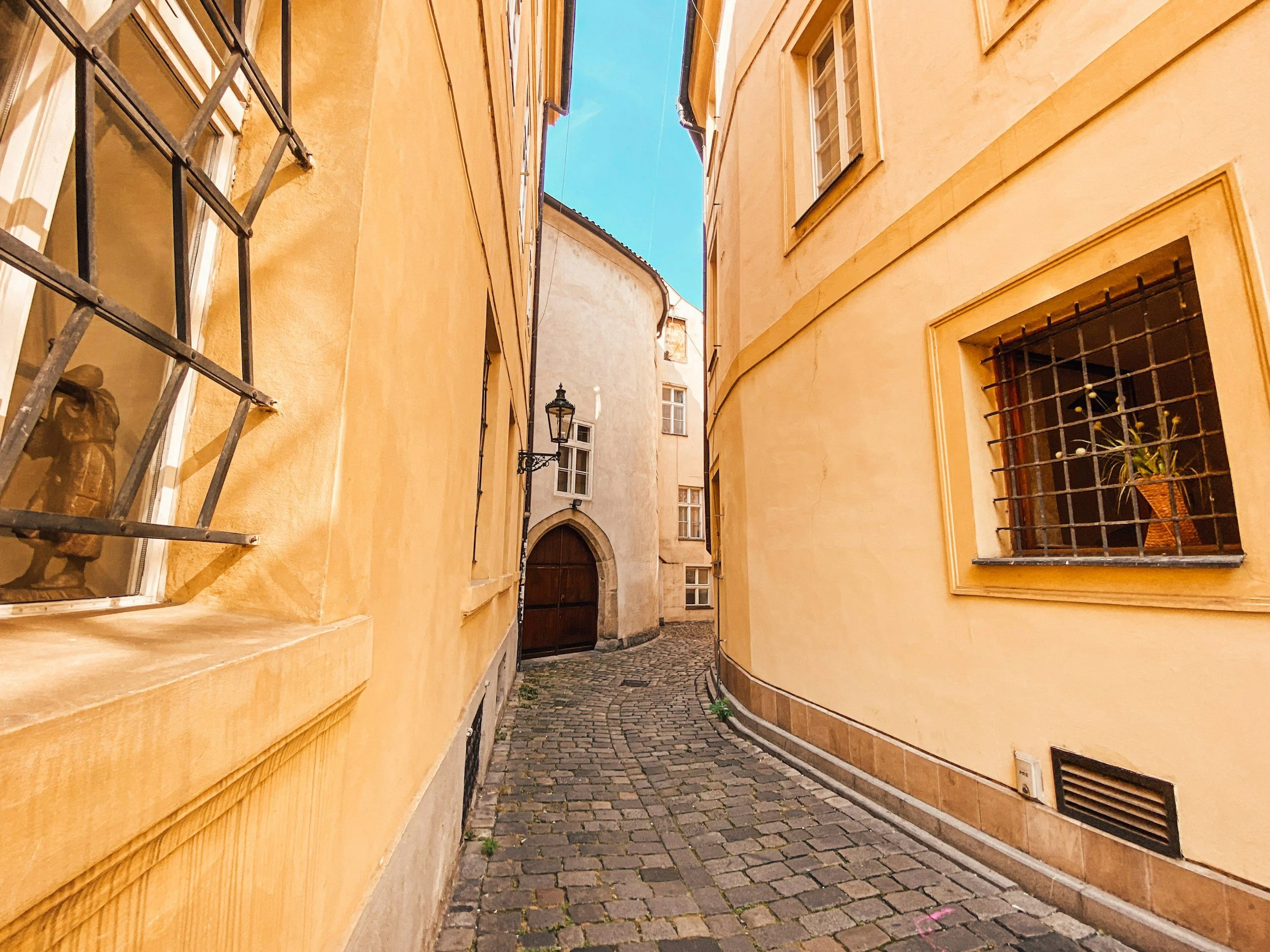 A narrow cobblestone alleyway between yellow and beige buildings with barred windows and a wooden door, under a clear blue sky.