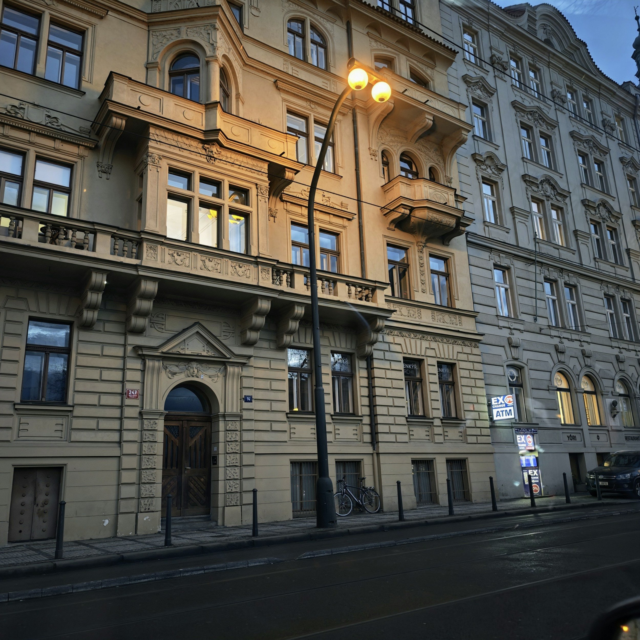 A European-style building with ornate architectural details, large windows, balconies, and a wooden entrance door on an urban street at dusk. There is a street lamp and a bicycle parked outside.