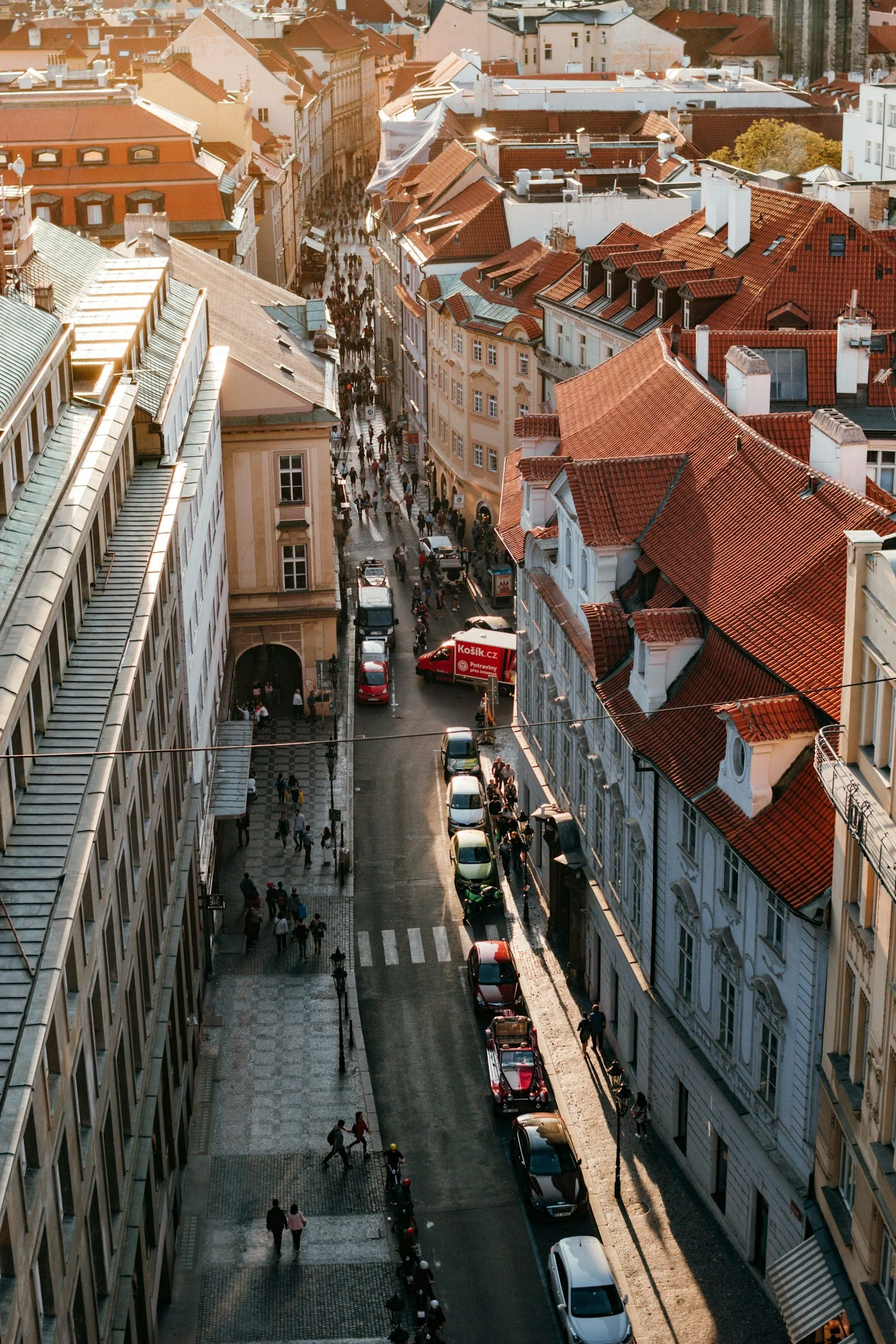 Aerial view of a busy city street with people walking, cars parked along one side, and historic buildings with red-tiled roofs.