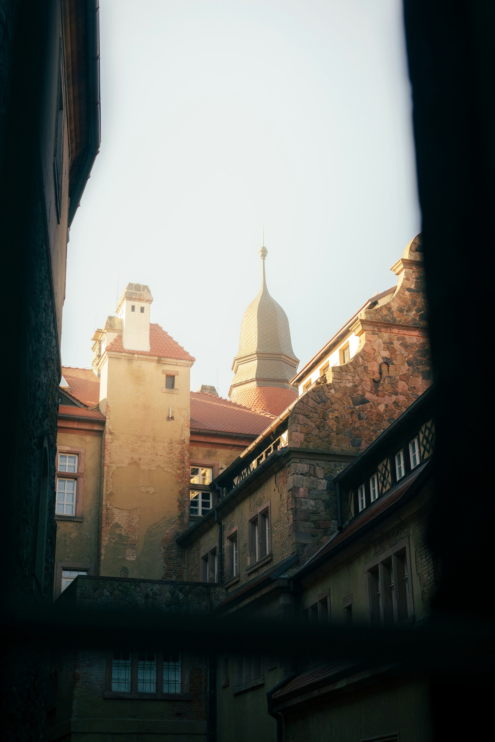 View of rooftops and a spire with a weather vane behind a narrow opening between buildings.