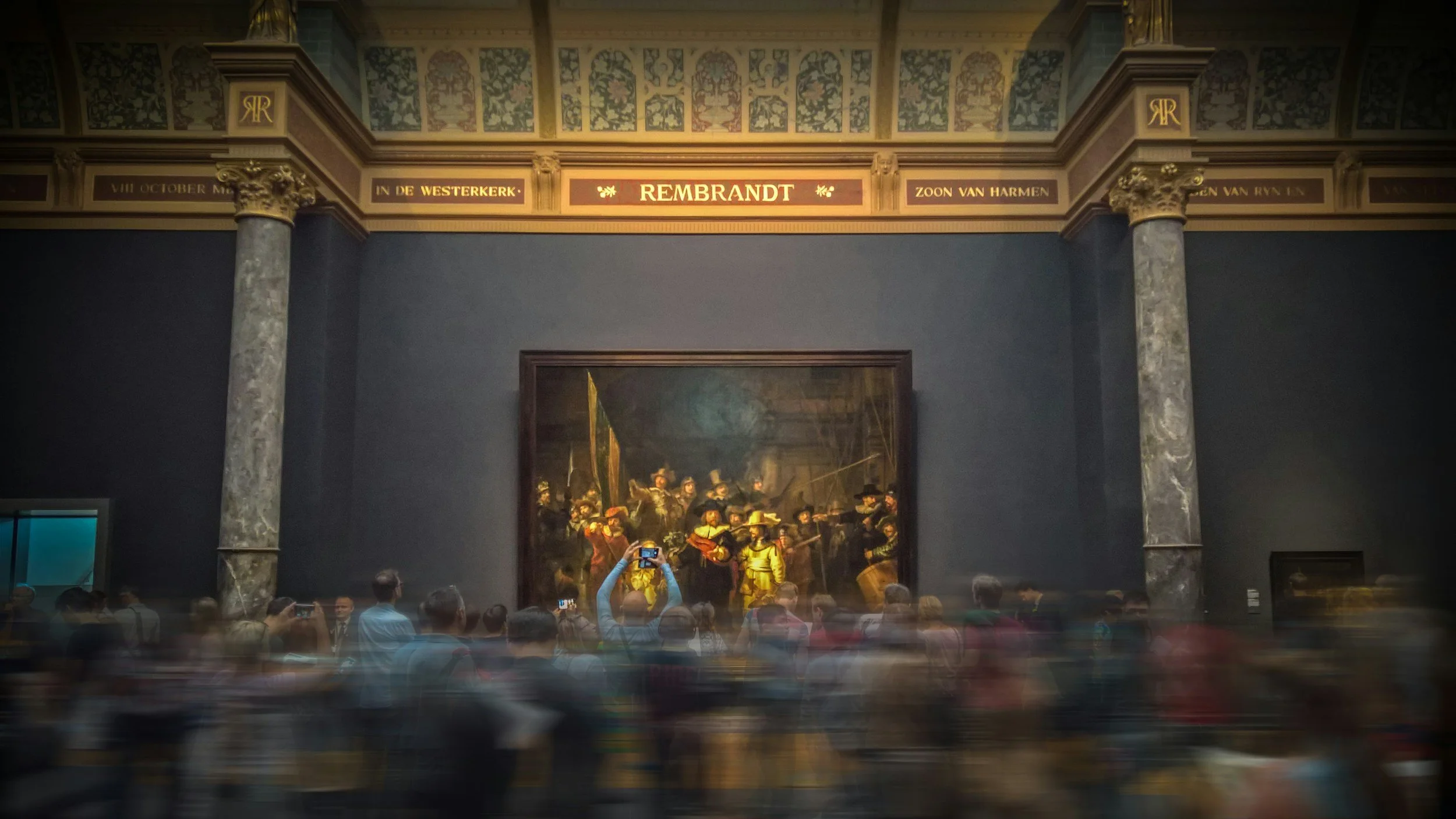 People viewing and photographing a large painting in an art gallery, with ornate architectural details and gold accents.