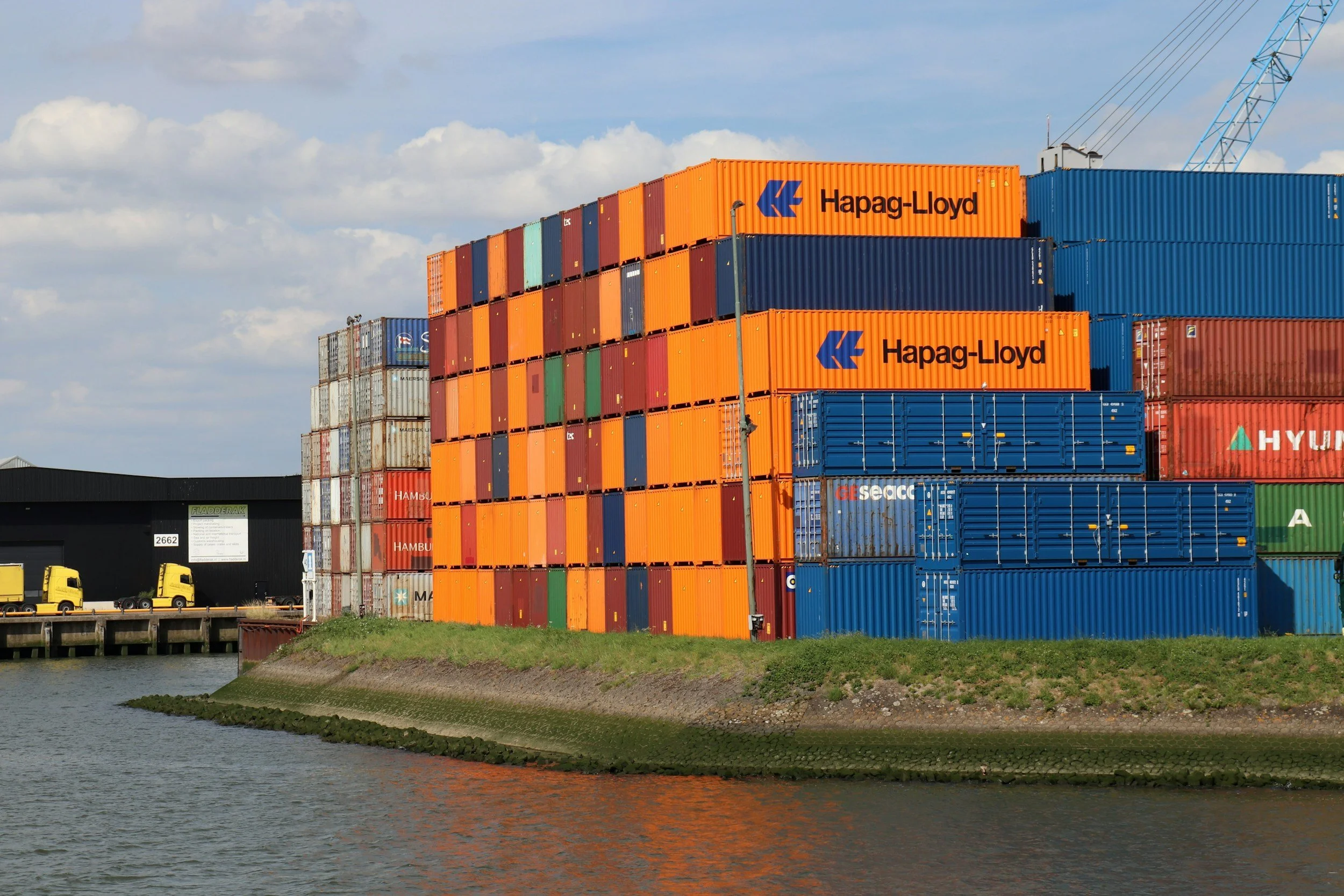 Stack of colorful shipping containers on a dock by a body of water under a partly cloudy sky.