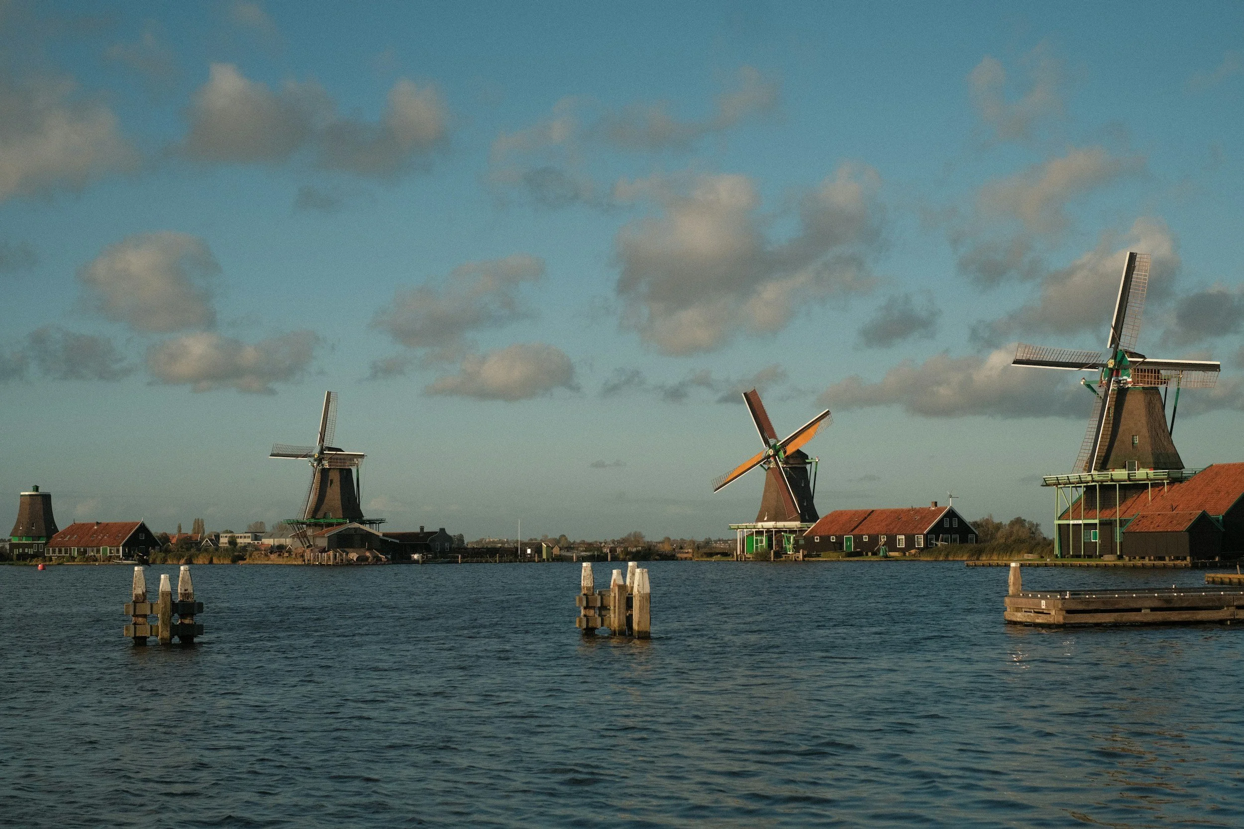 Three traditional Dutch windmills beside a body of water with wooden dock posts, under a partly cloudy sky.