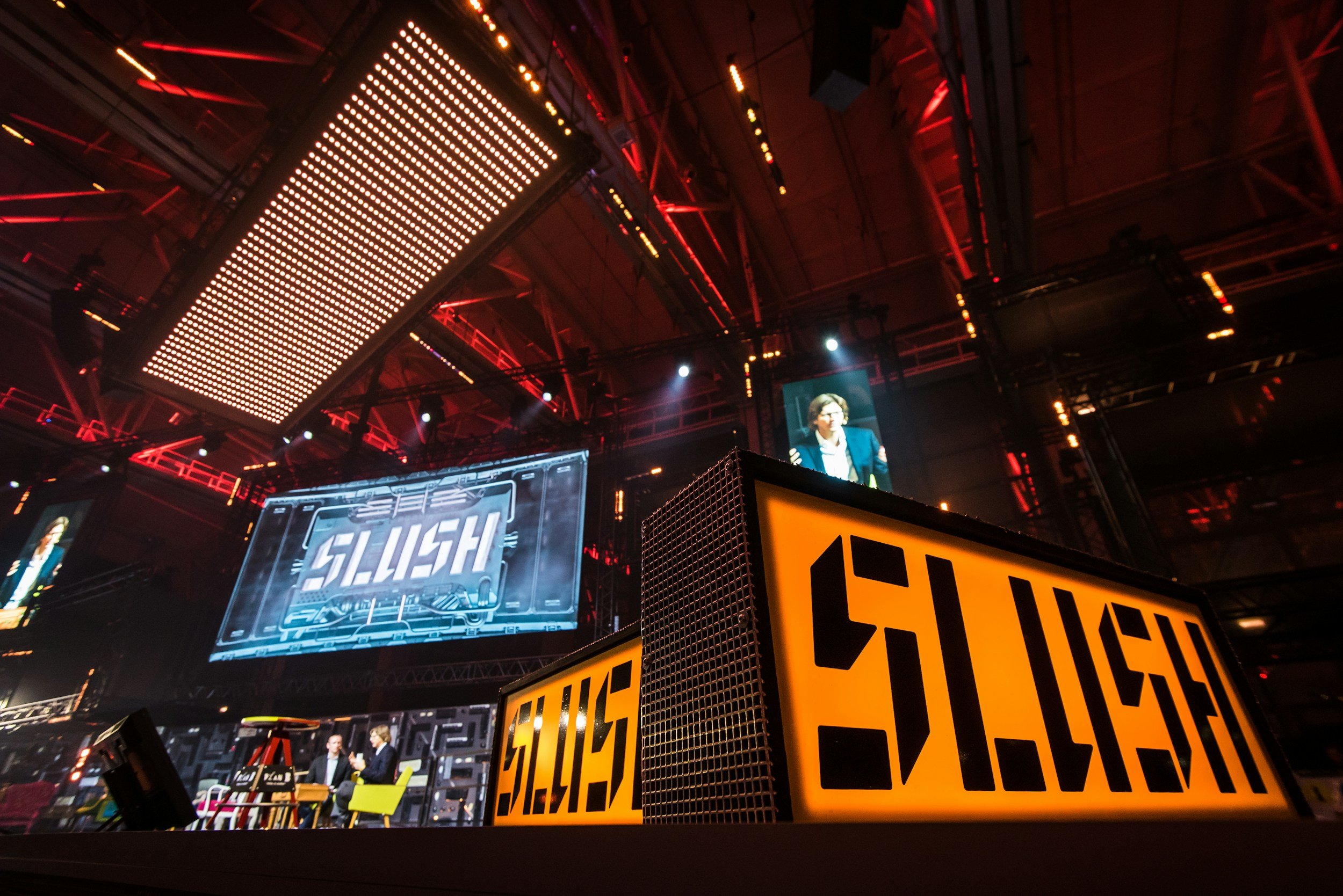 Stage at a conference or event with large illuminated signs that read 'SLUSH', two people seated and two screens displaying the same 'SLUSH' logo, and red lighting overhead.