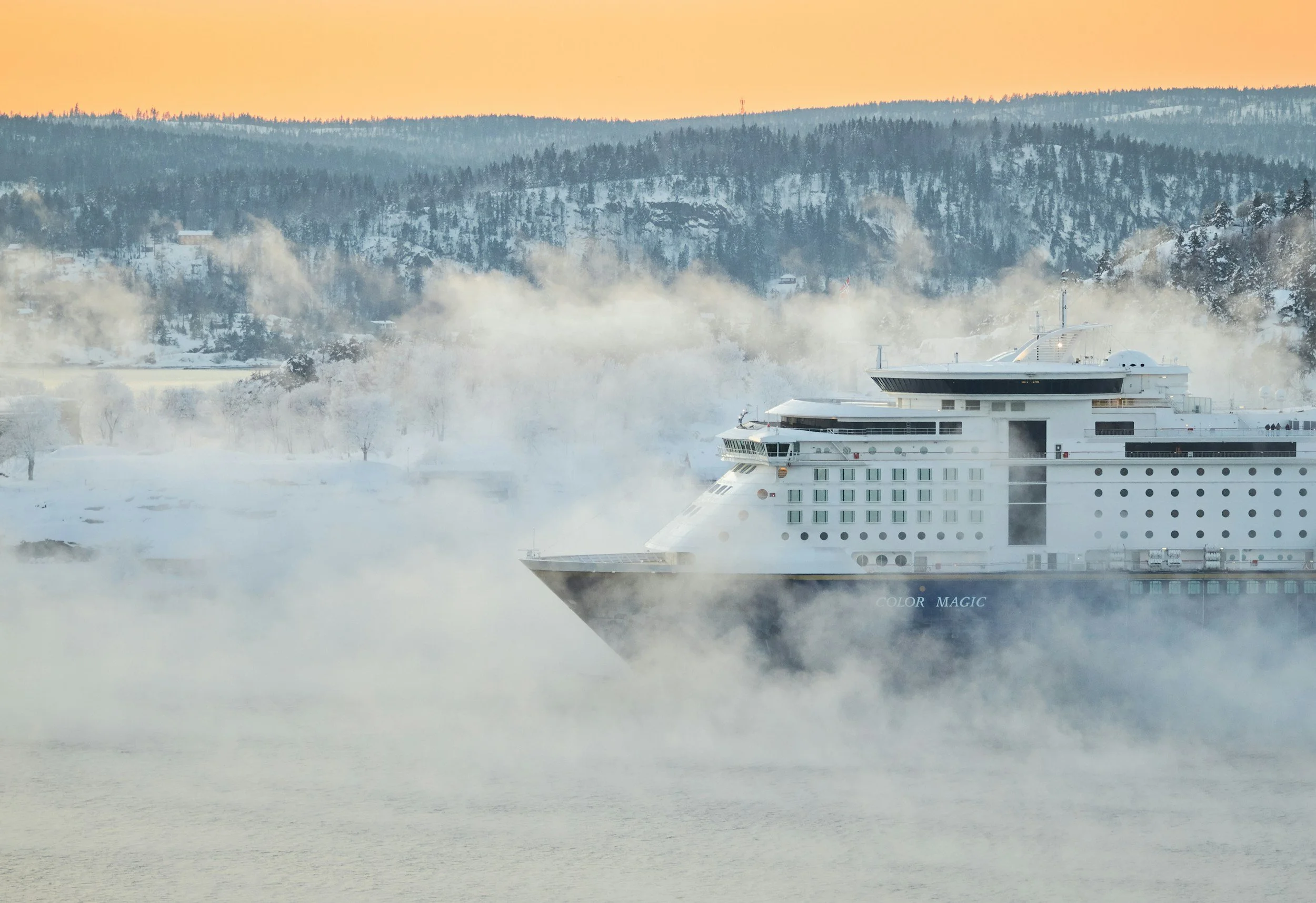 A large white cruise ship named 'Color Magic' sailing through icy water with snow-covered hills and trees in the background, surrounded by mist or steam near the water's surface.