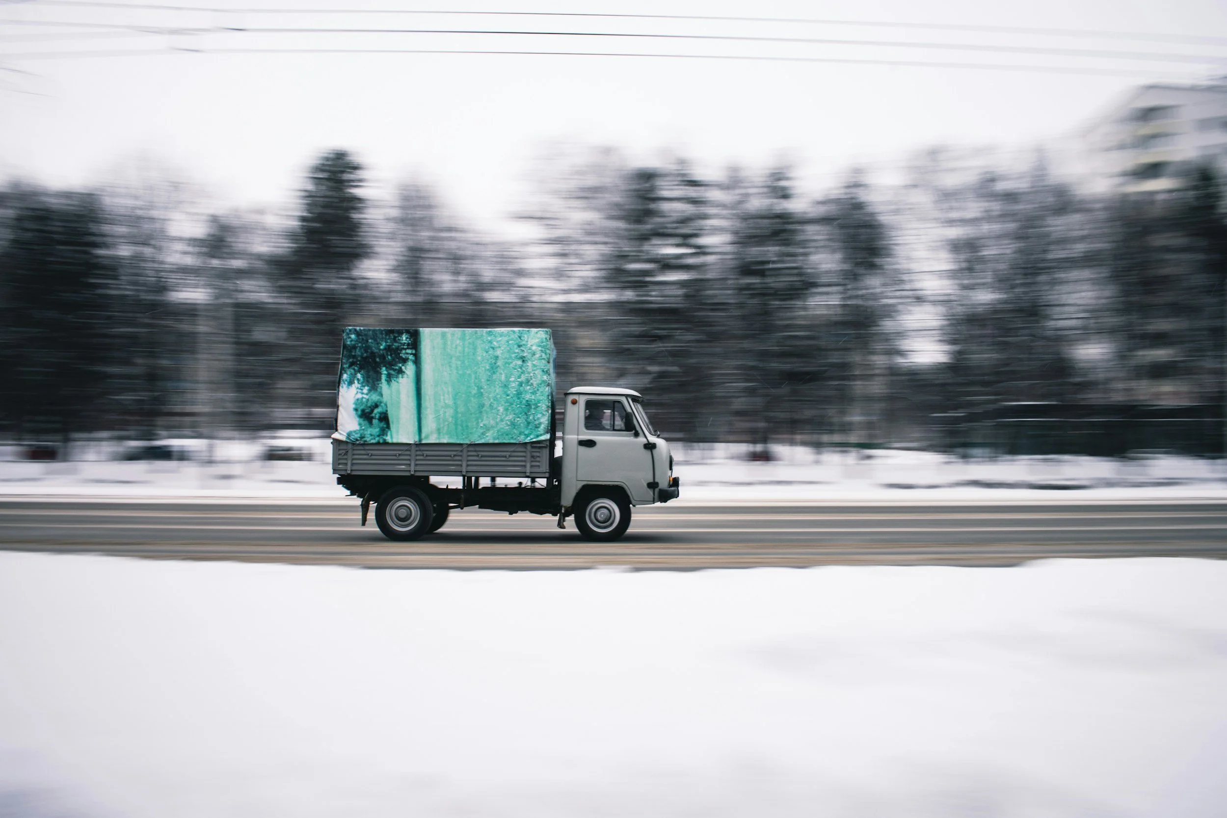 A small gray truck driving on a snowy road with a blurred forest background.