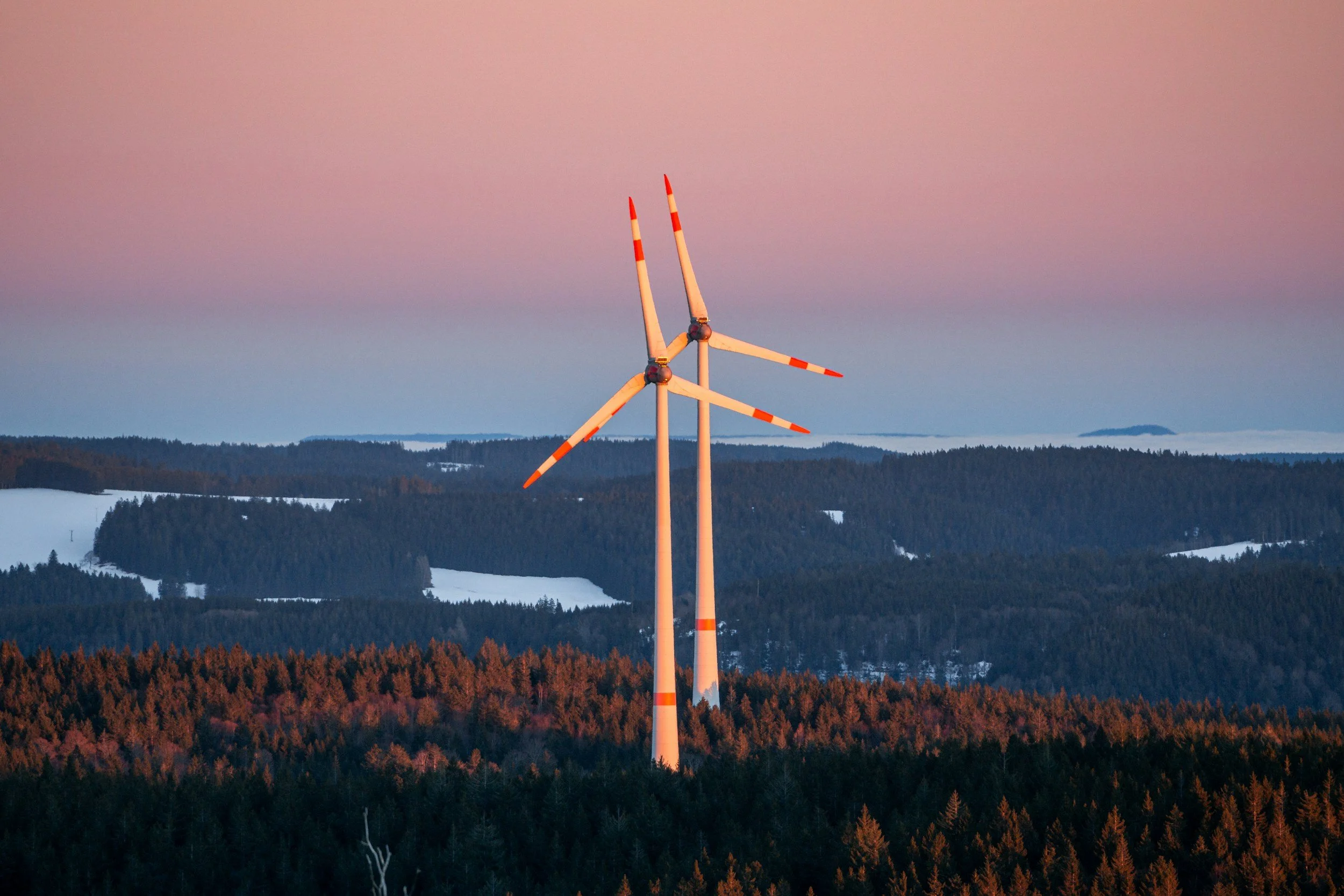 Three wind turbines on a hilltop at sunset, with a purple sky and forested landscape in the background.