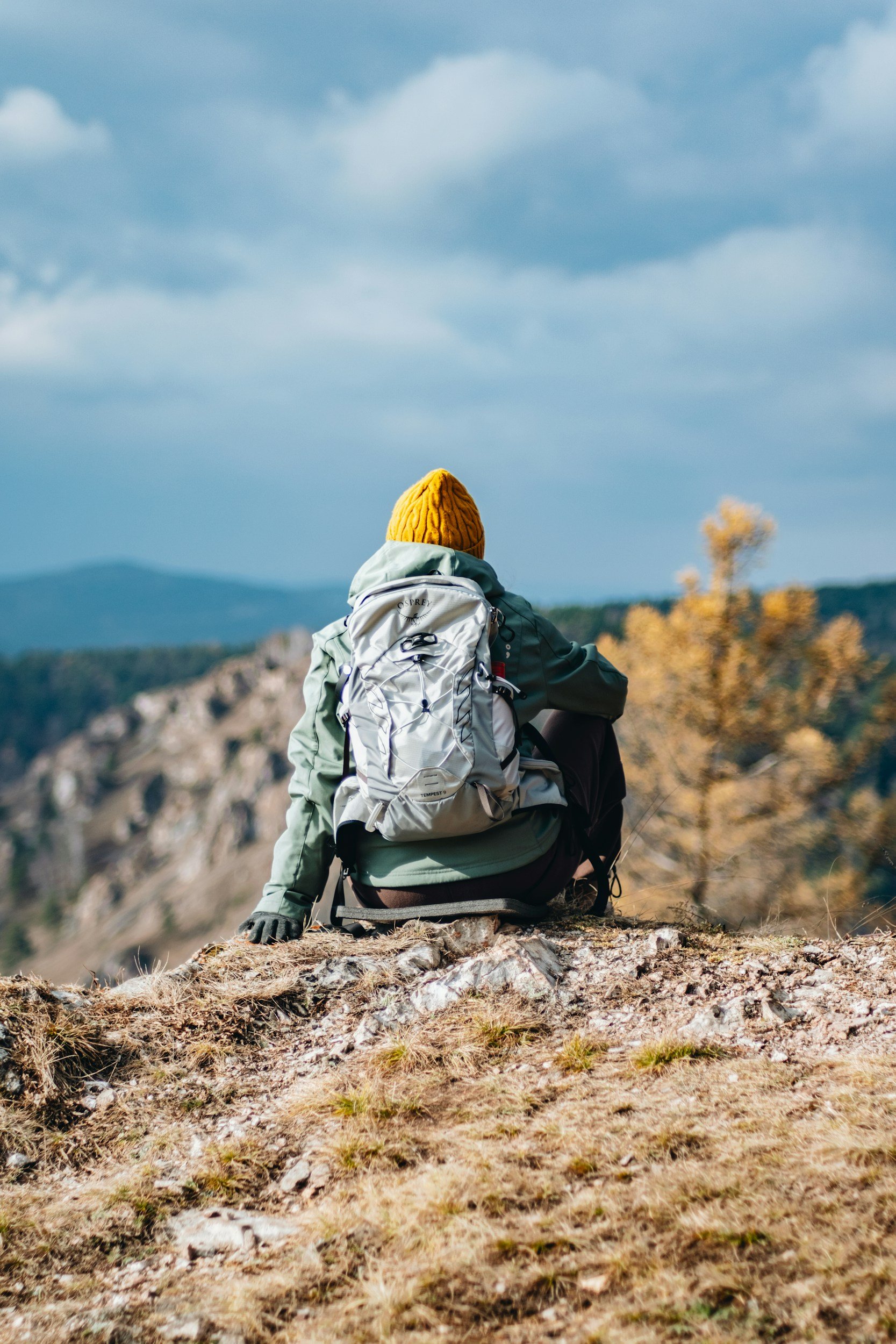 Person sitting on rocky ground outdoors, wearing a yellow knit cap and backpack, overlooking a landscape of hills and trees with cloudy sky.