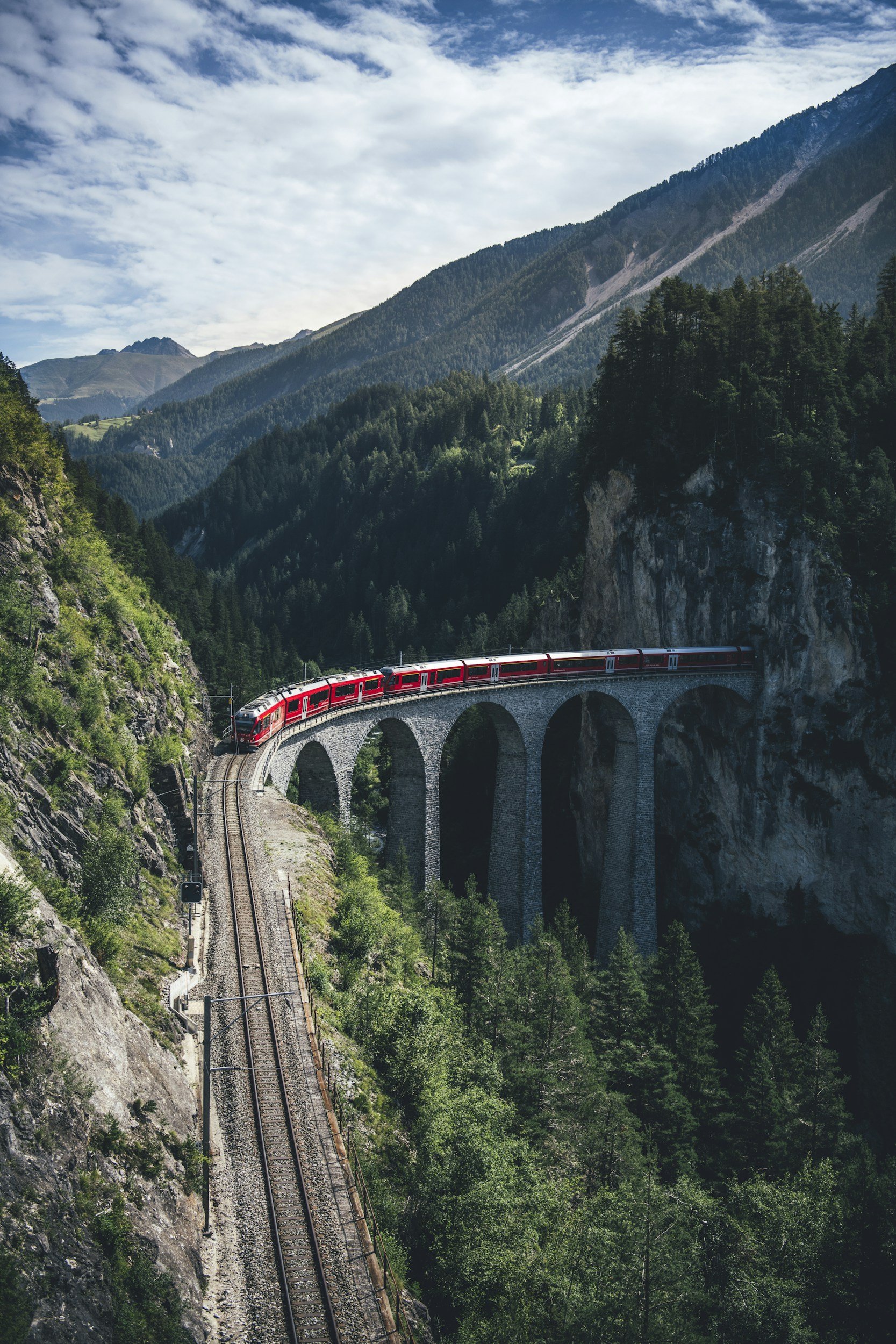Red train traveling over a high stone arch bridge in a mountainous forest landscape with a partly cloudy sky.