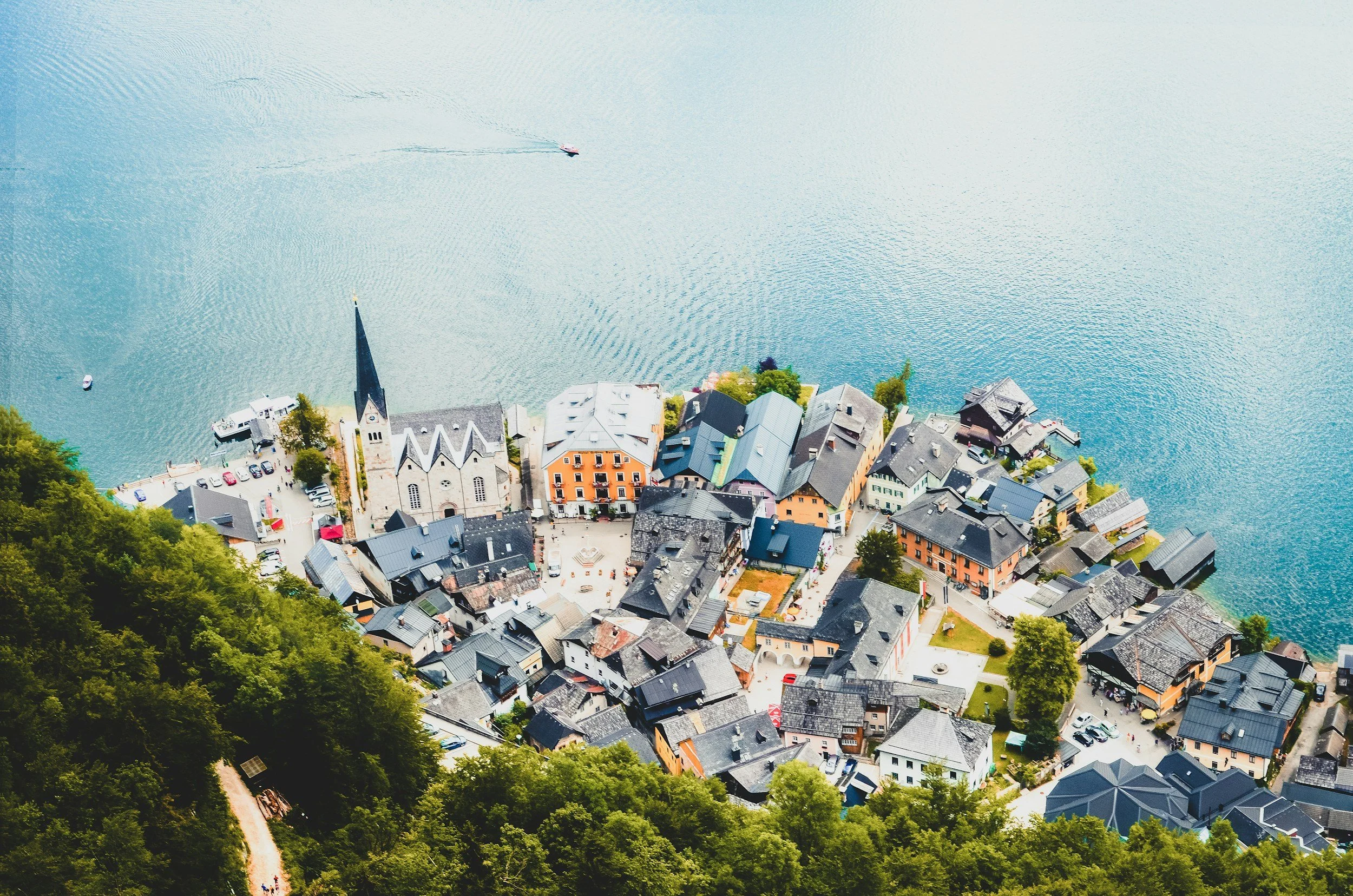 Aerial view of a small lakeside town with colorful buildings, a church with a tall steeple, and a forested hillside in the foreground, next to a large body of water with boats.