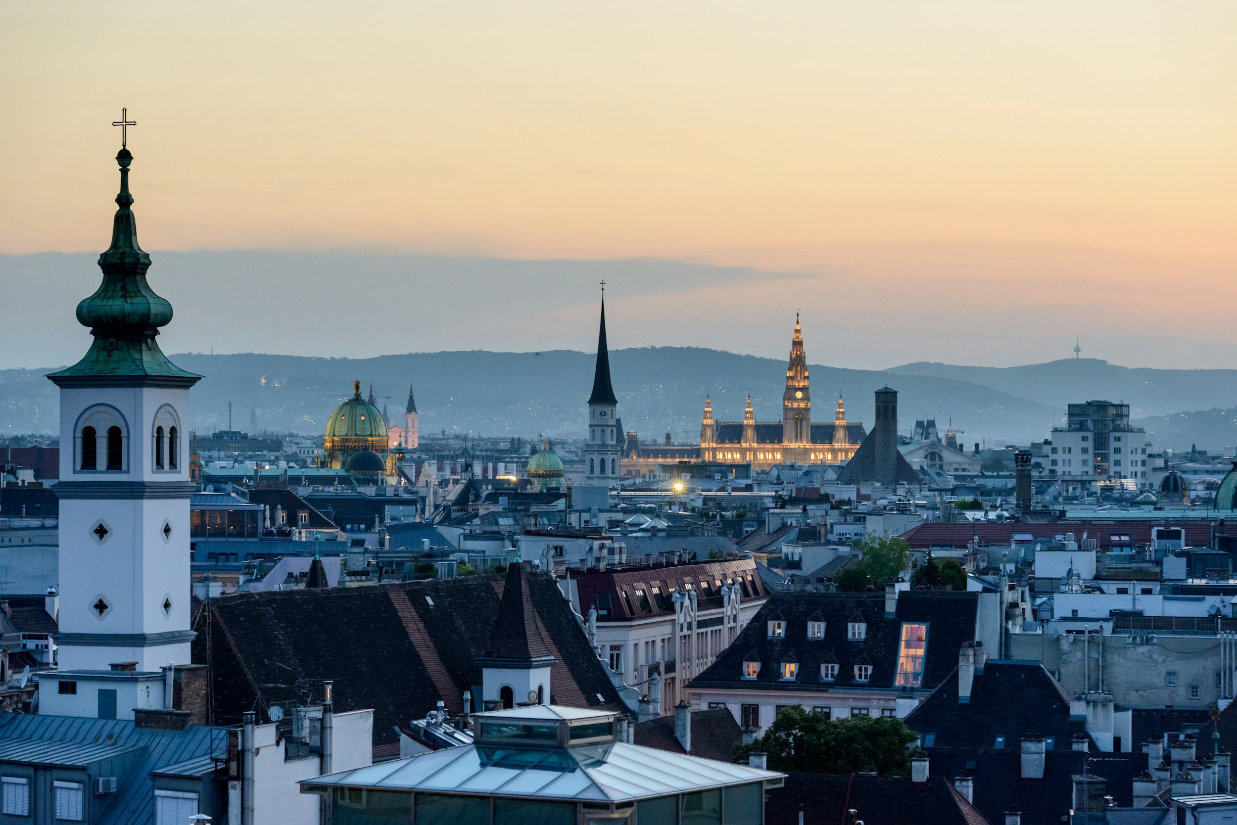 A city skyline at dusk with church steeples, domes, and historic buildings, featuring a mountain range in the background.