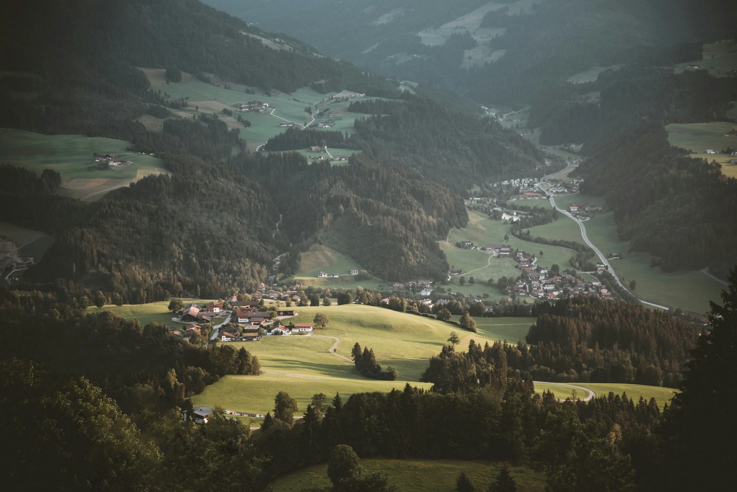Landscape view of a lush green valley with scattered houses, forests, and winding roads in rolling hills and mountains in the background.