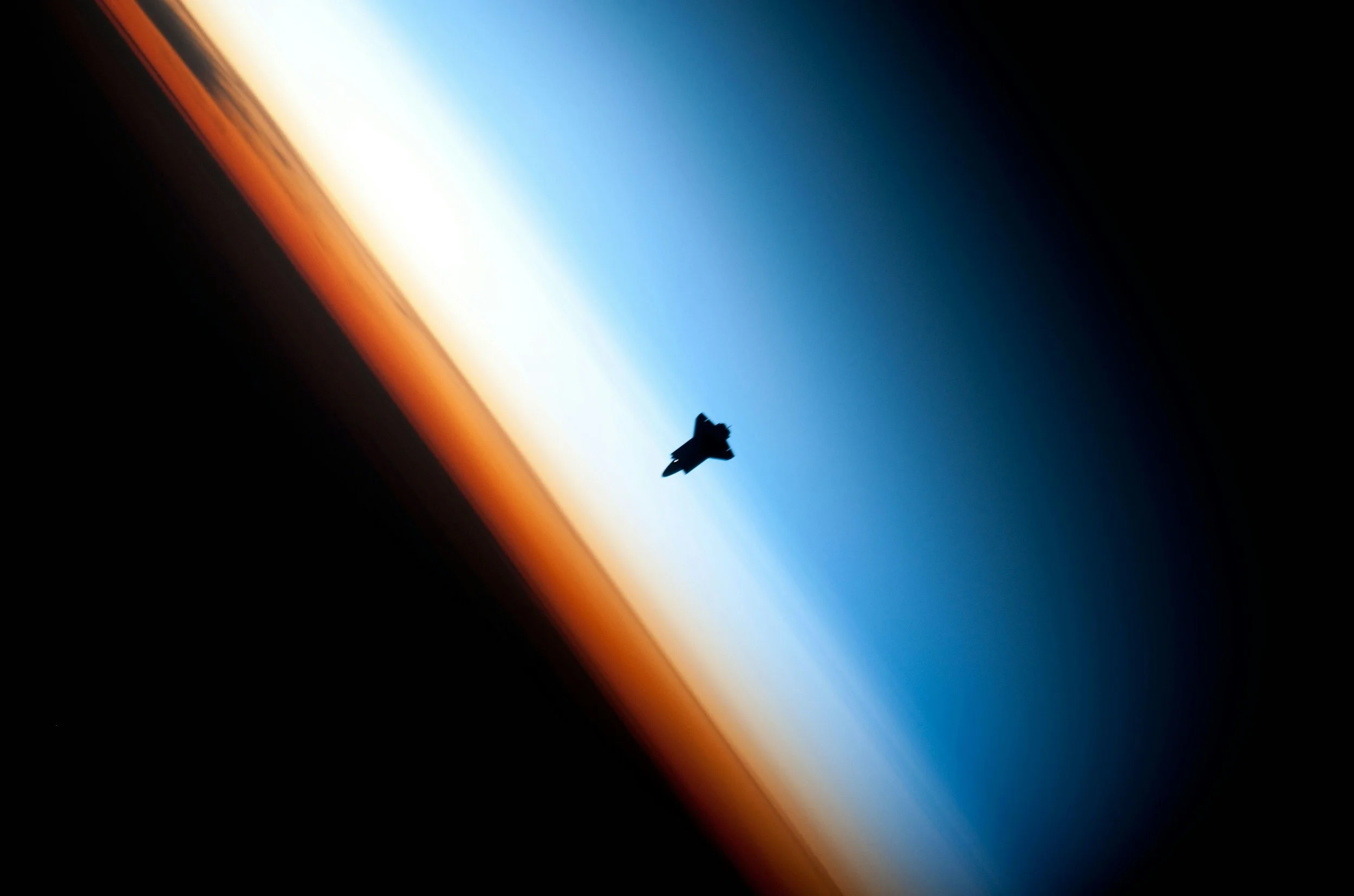 Photo of a space shuttle orbiting Earth during sunrise, with the planet's curved horizon showing layers of orange, white, and blue atmosphere.