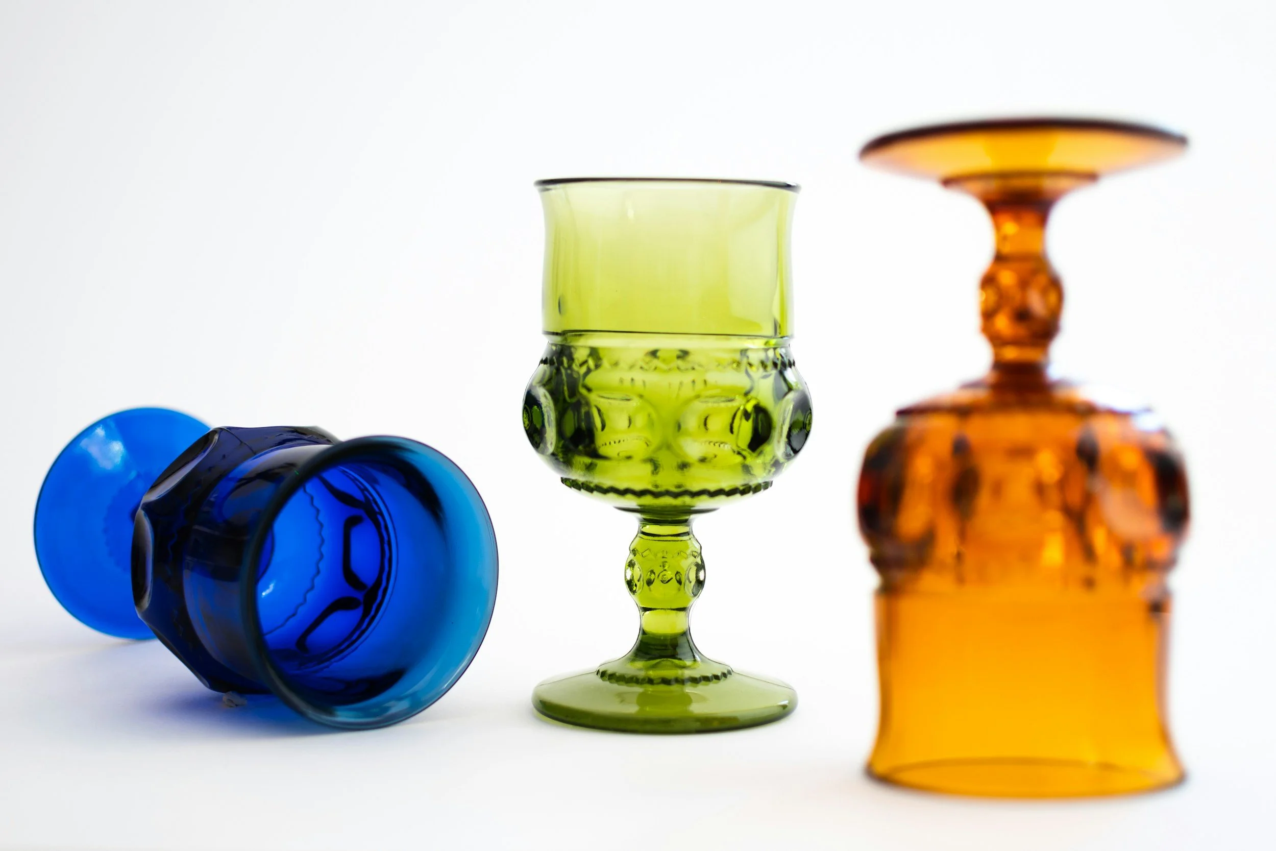 Four colorful glass objects: a fallen blue glass, a green glass goblet, and an orange glass vase on a white background.