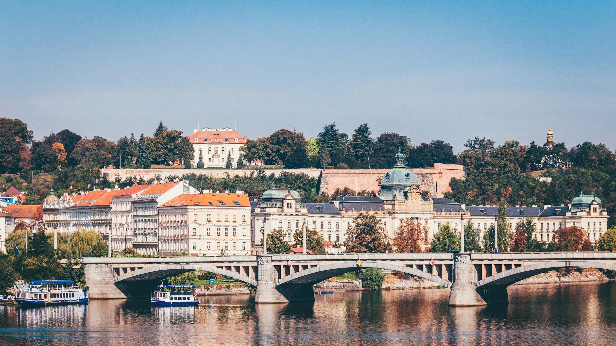 Scenic view of a city with historic buildings, a bridge over a river, and a hill with trees and a large mansion in the background.